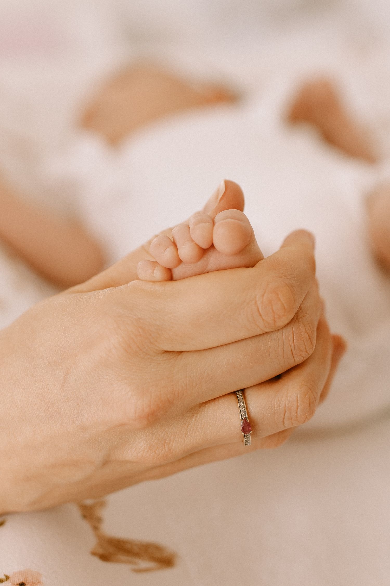 Adult hand gently holding tiny baby foot with delicate ring on finger against soft white background.