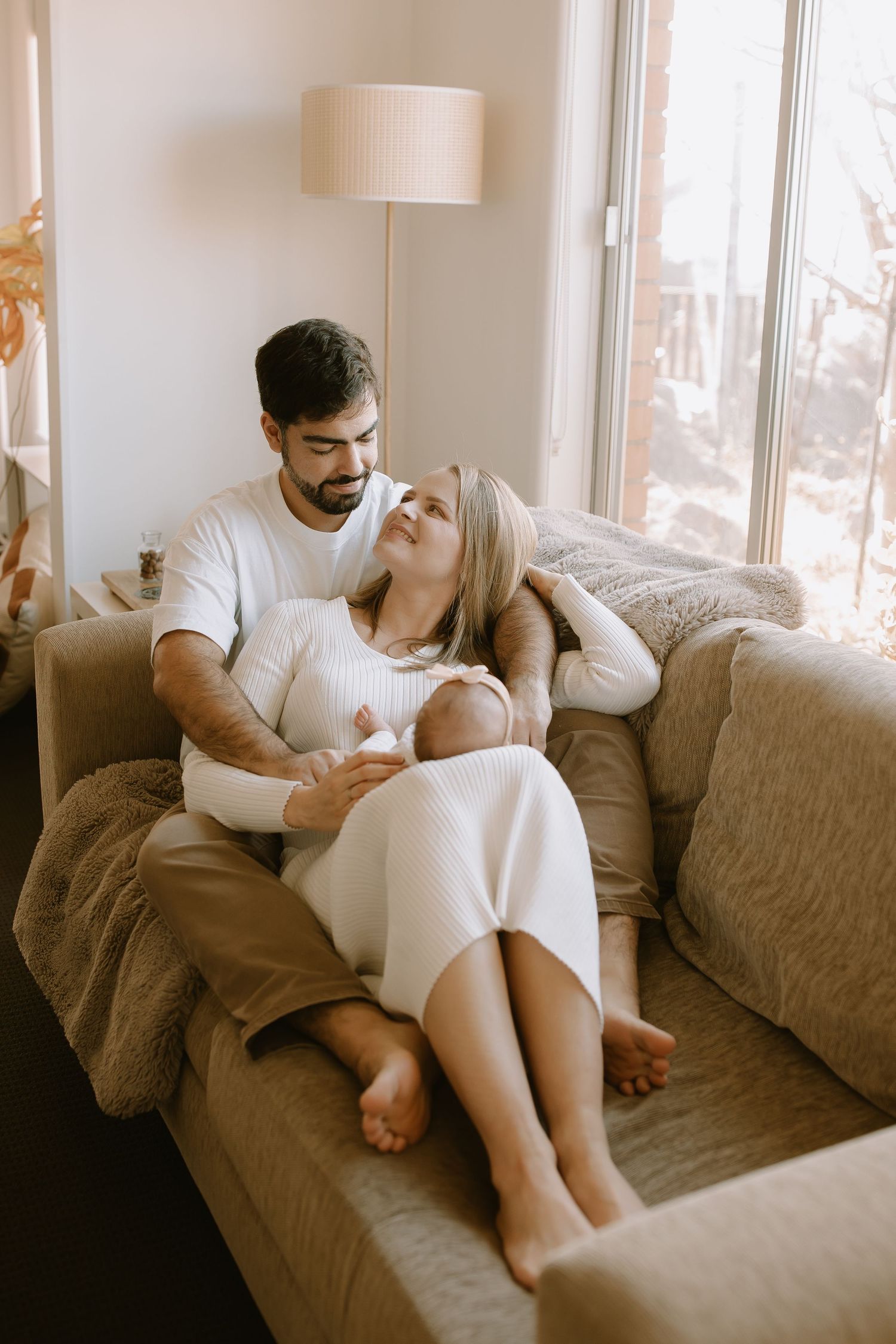 Tender romantic moment shared between couple relaxing and cuddling together on brown couch near window with floor lamp.