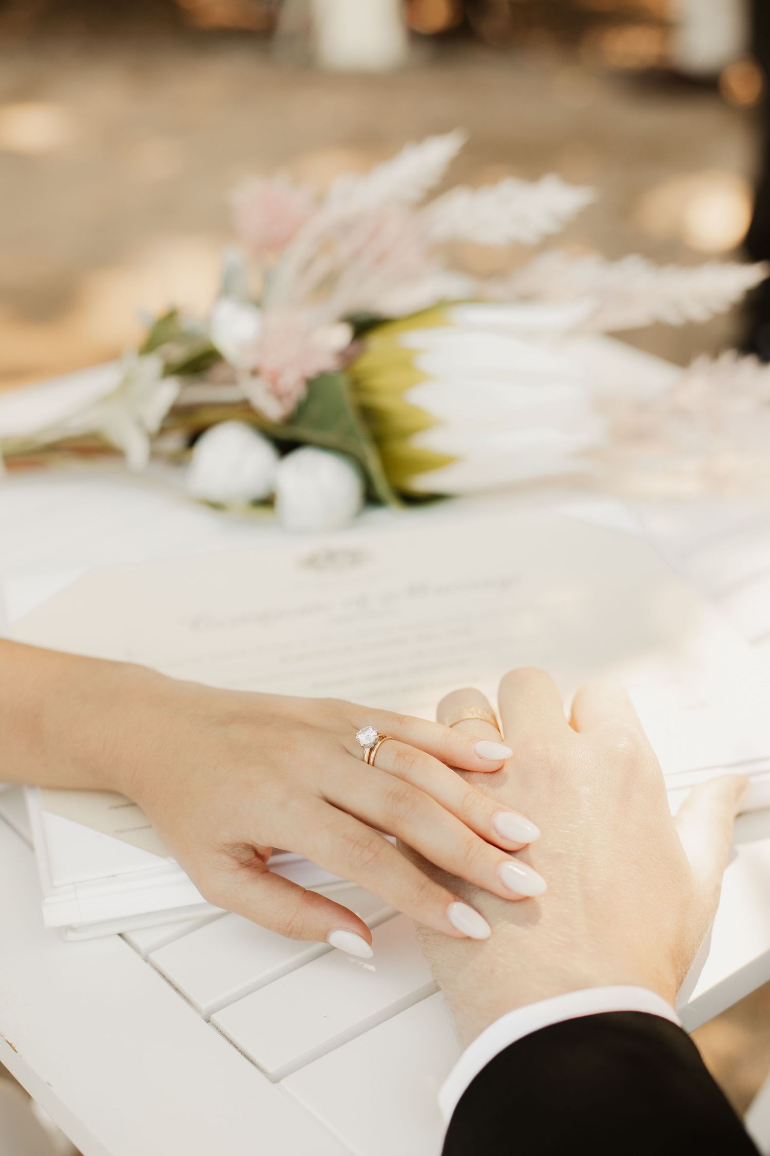 Close-up of hands with wedding rings overlapping on a white surface with soft pink flowers in the background.