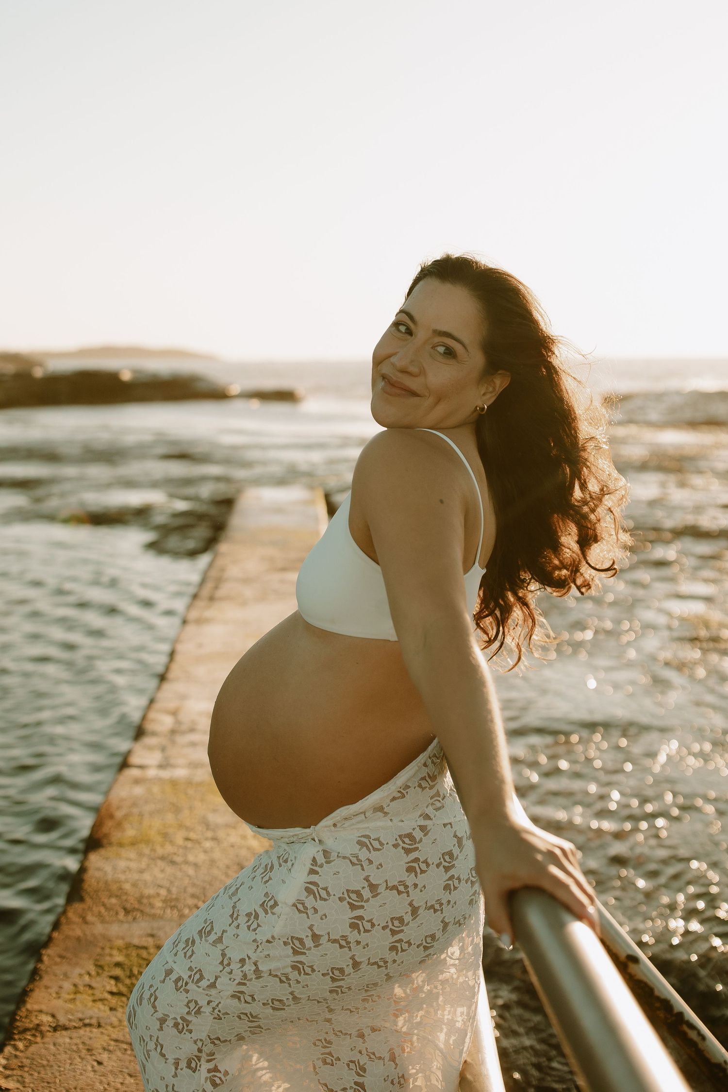 Expectant mother in white lace dress poses by ocean during sunset maternity photoshoot.