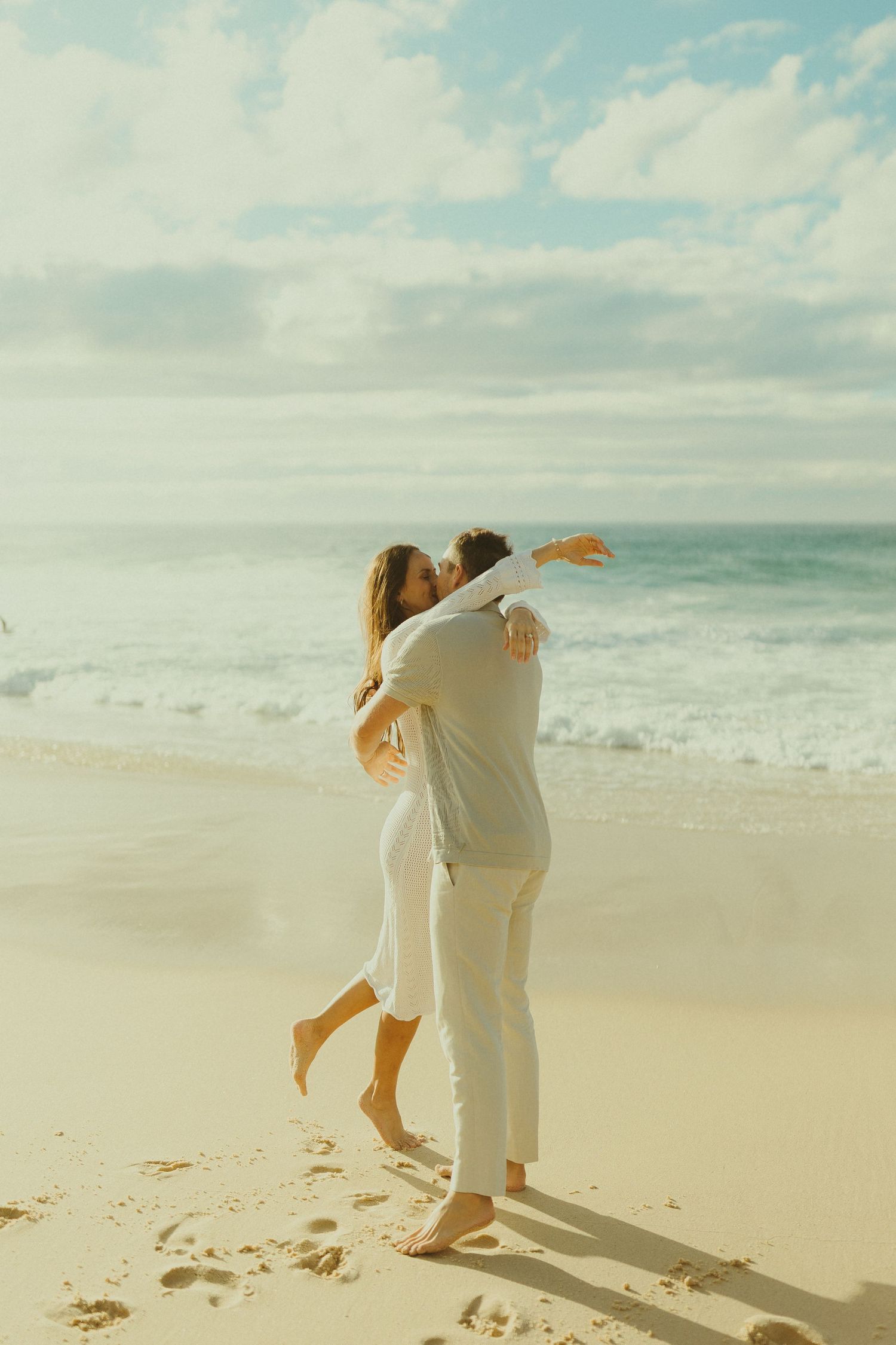 A romantic couple in light colored clothing embracing and dancing on a sunny beach.