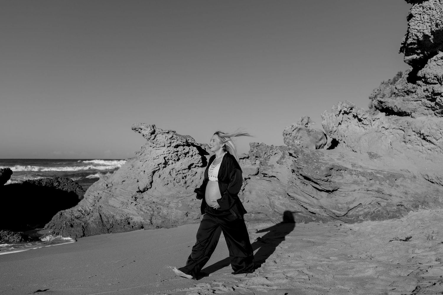 Black and white photo of a person jumping joyfully across rock formations at a beach coastline.