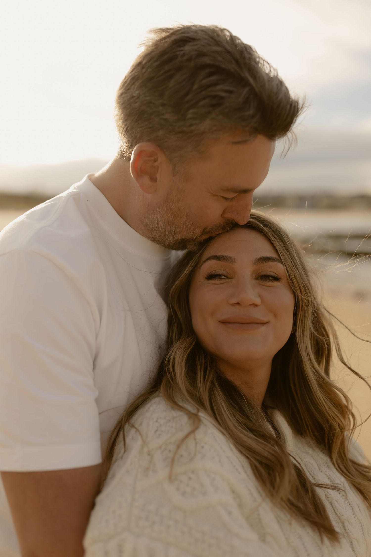 Man gently kisses smiling pregnant womans head on beach at golden sunset, tender maternity moment.