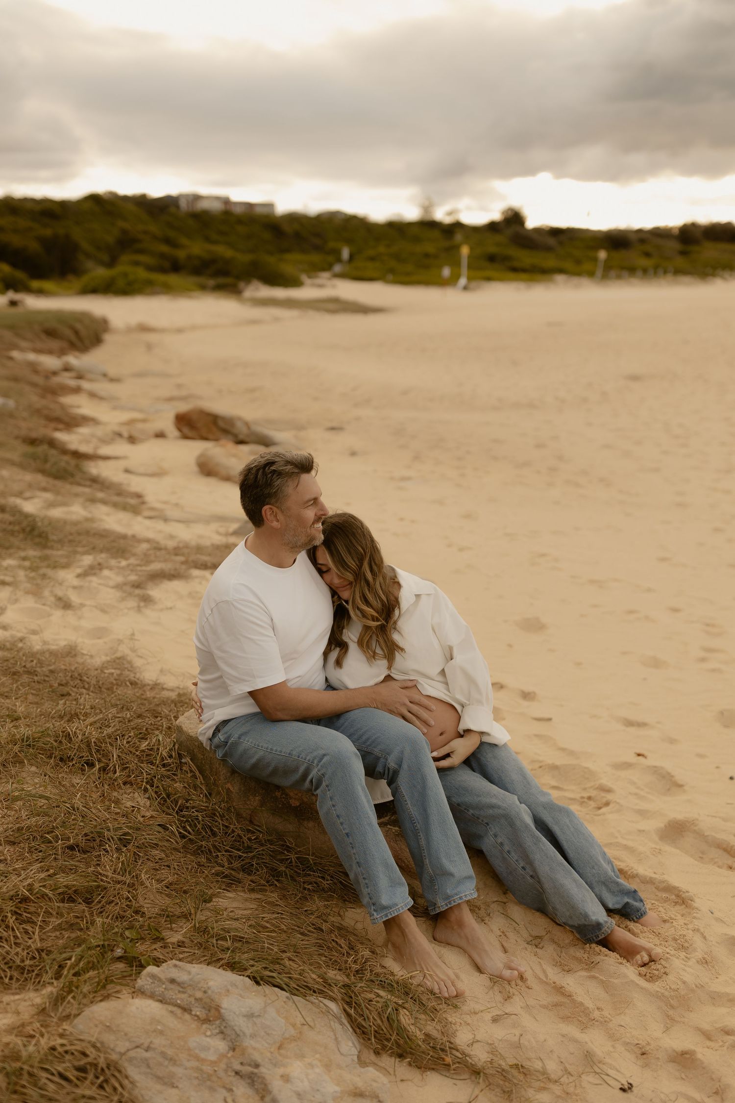 Couple sitting on sandy beach dunes embracing, maternity photo shoot at sunset.