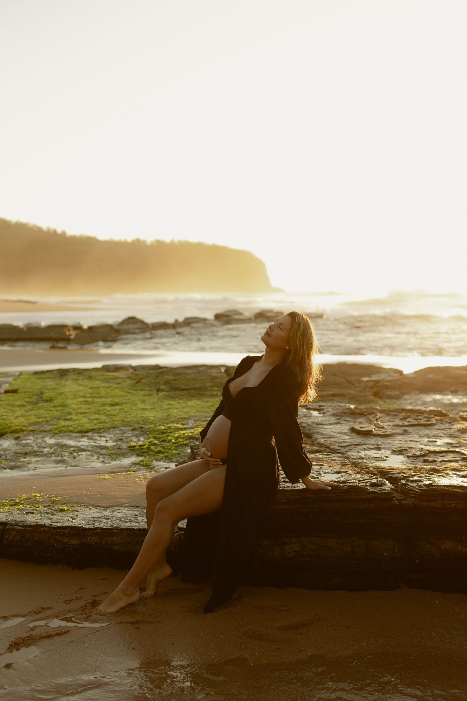 Woman in black dress sitting on driftwood at sunset beach, golden light illuminating rocky shoreline and ocean waves.