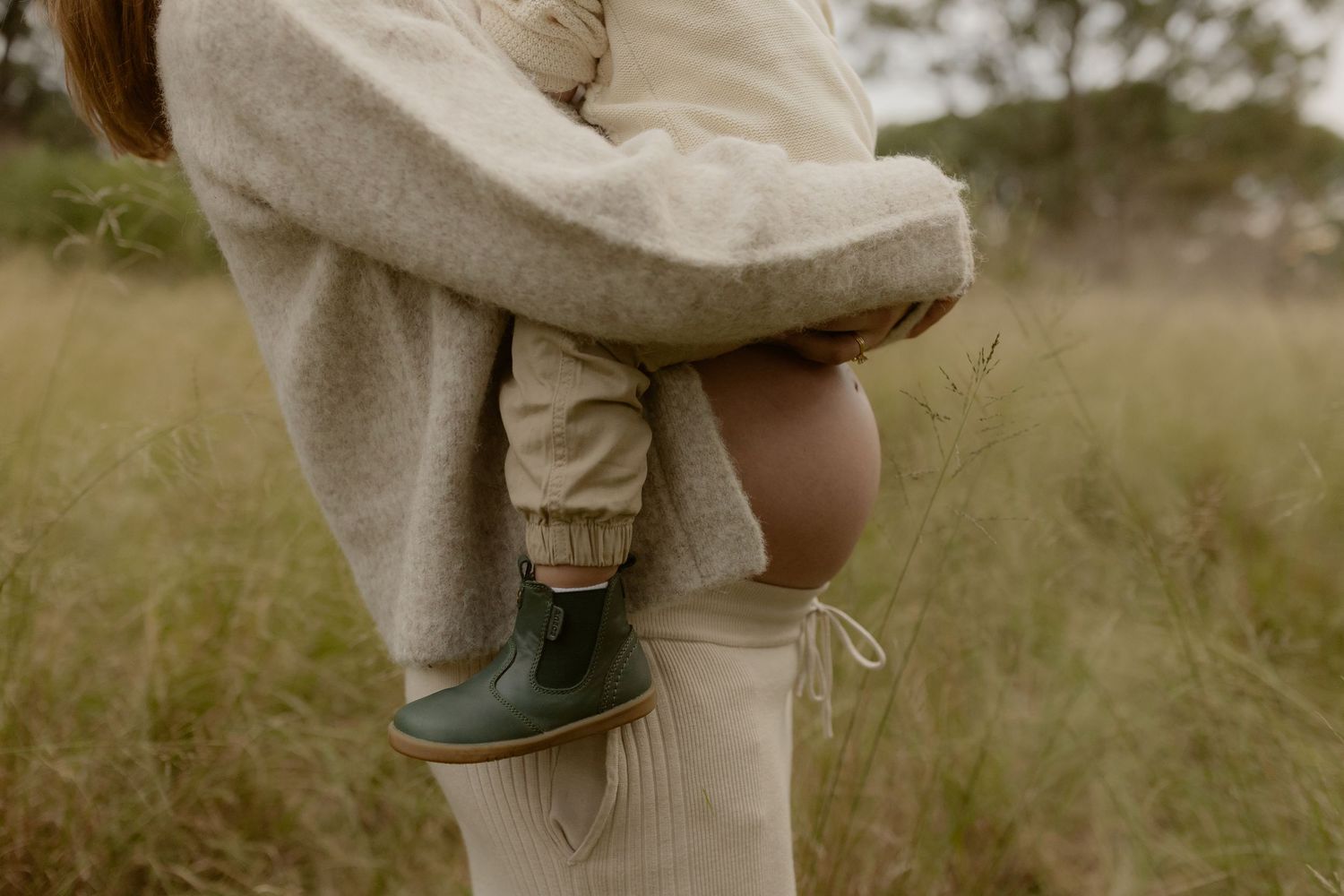 Close-up of pregnant woman in cream outfit holding toddler wearing green boots in grassy field.