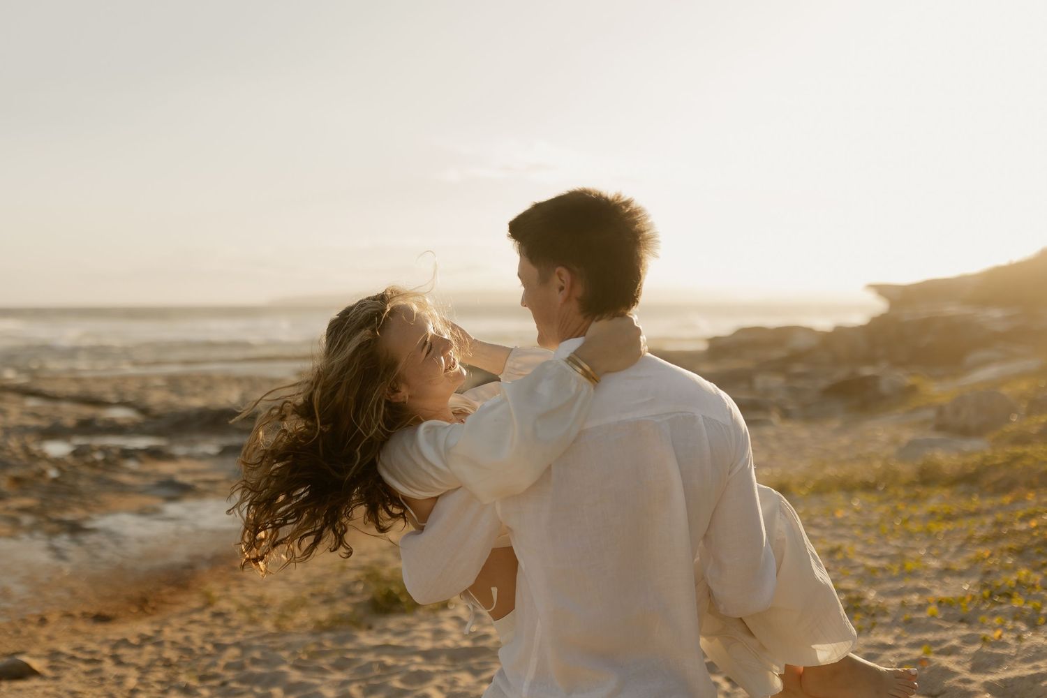 Couple in white clothing embracing during sunset beach photography session.