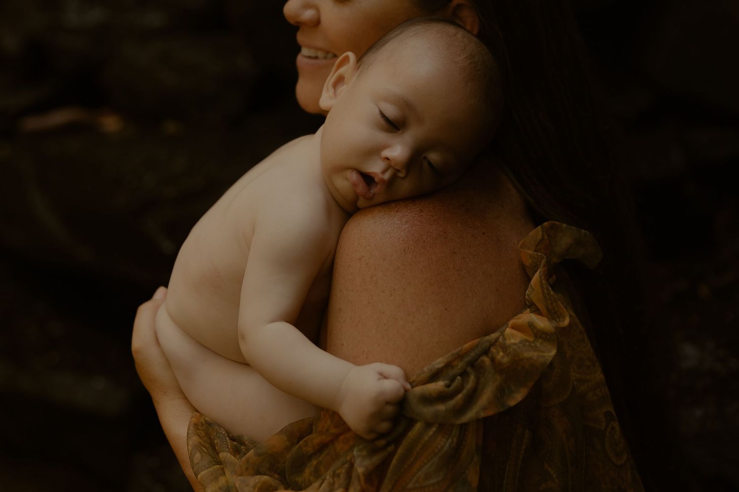 A sleeping baby rests peacefully against a caregiver's shoulder in a warm, moody lighting.