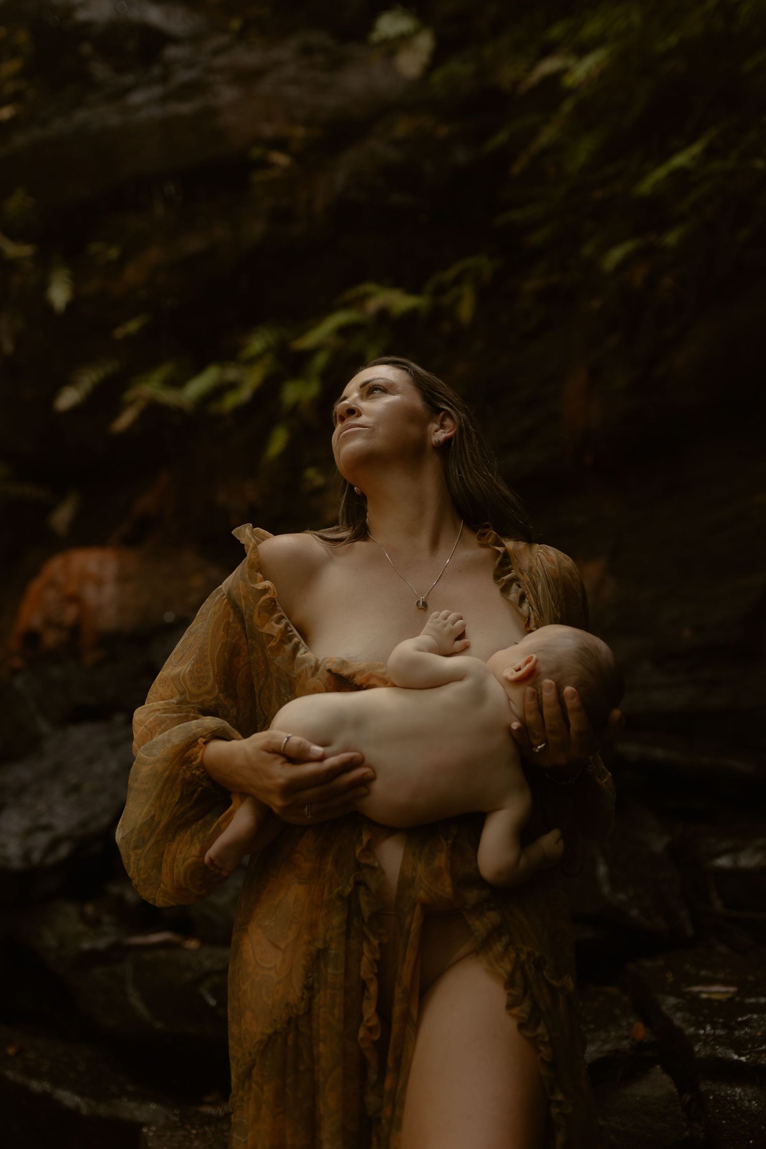 A tender moment captured in moody lighting shows a figure in brown dress cradling an infant against dark rocky surroundings.