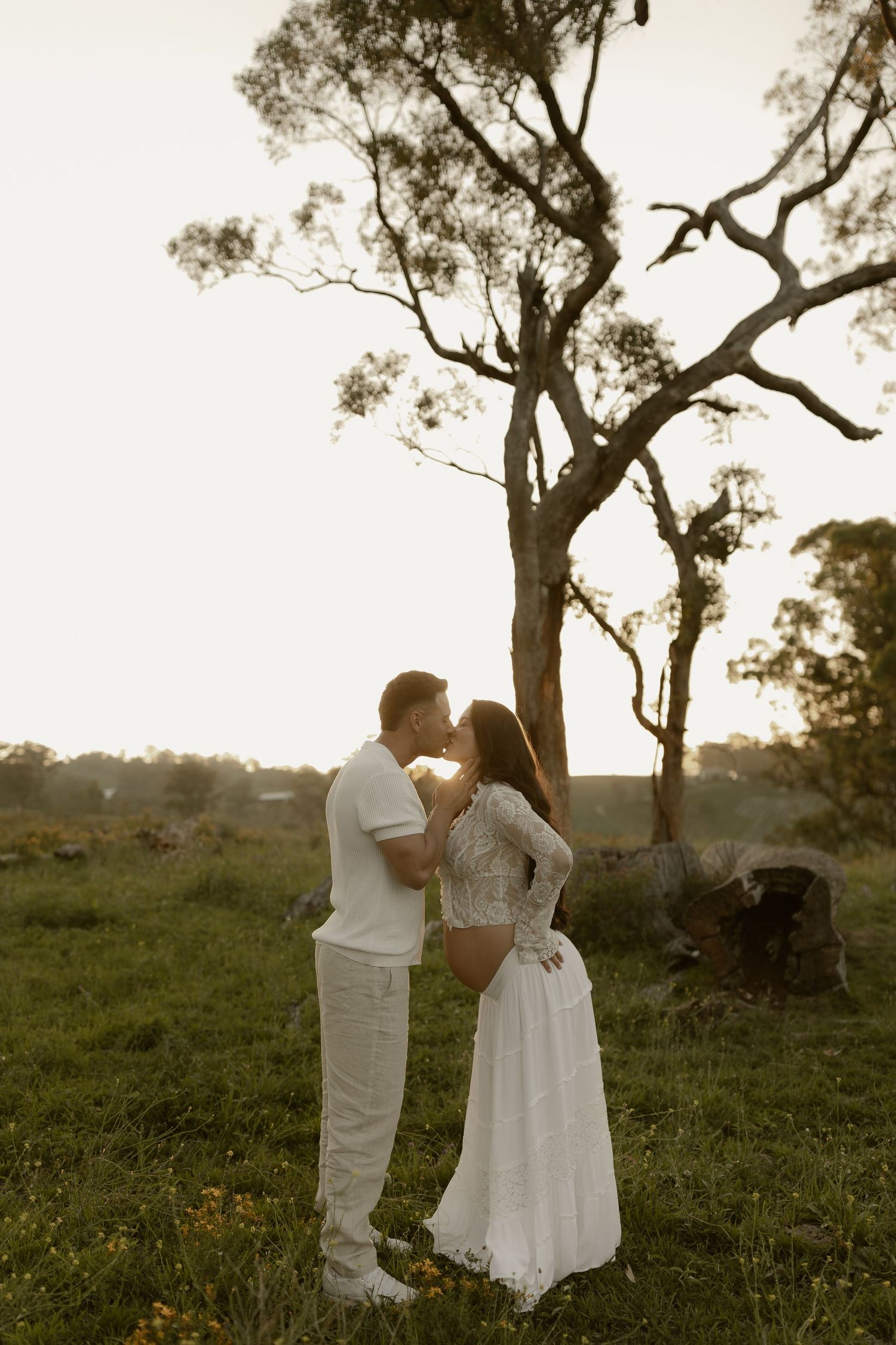 Couple in white outfits shares romantic moment under large tree at sunset.