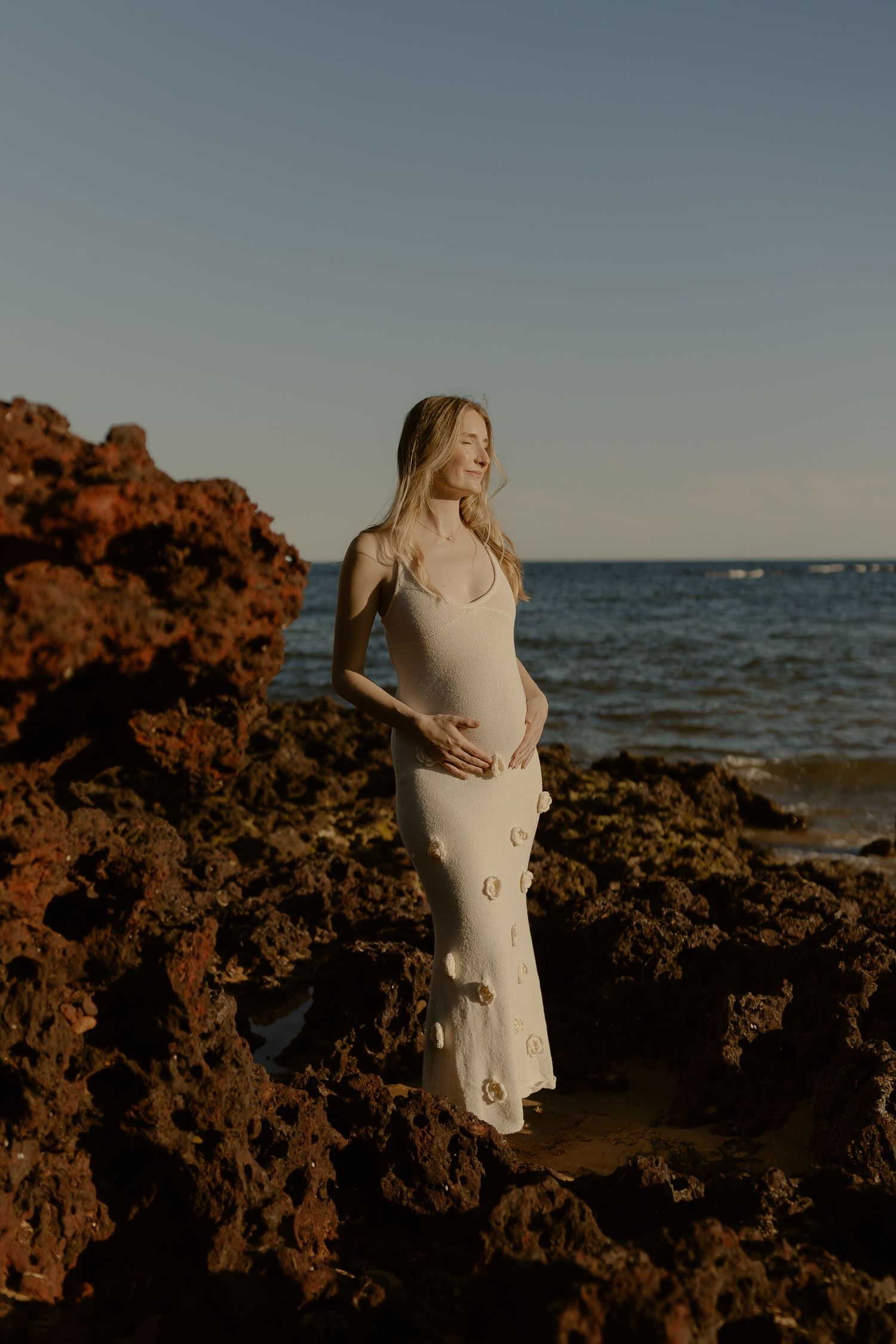 A dreamy maternity photoshoot on a rocky beach at sunset with warm golden light.