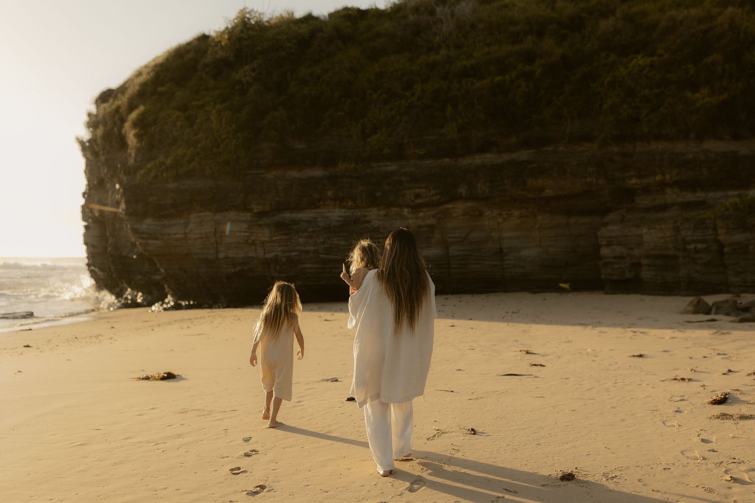 Two figures walk along a scenic beach with dramatic cliffs at sunset creating long shadows on the sand.