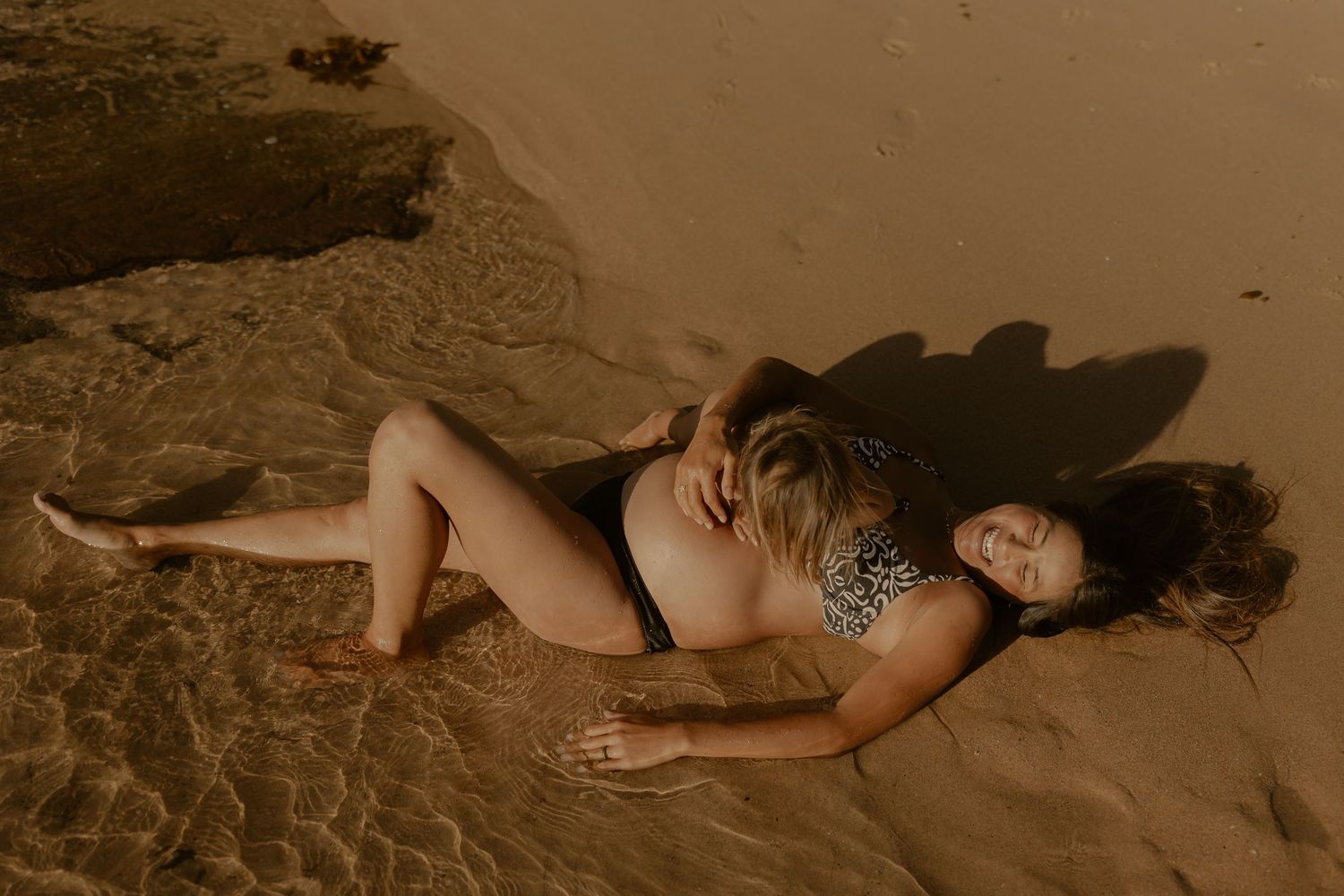 Pregnant woman in swimwear lying peacefully on sandy beach during sunset.