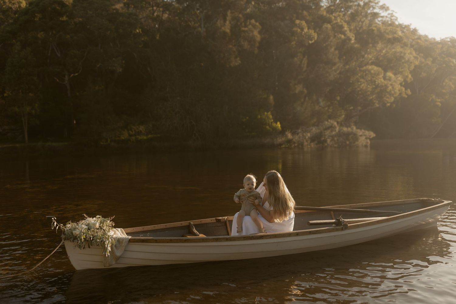 Two women in white dresses sit in a wooden rowboat on a calm sunlit lake surrounded by lush green trees.