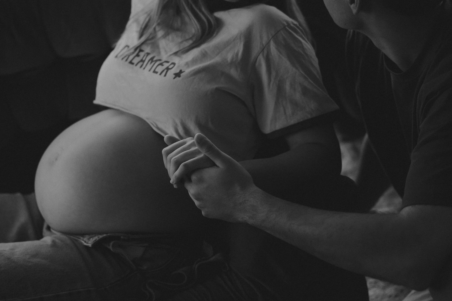 Black and white close-up of pregnant belly with partner gently holding sides, wearing Dreamer shirt.