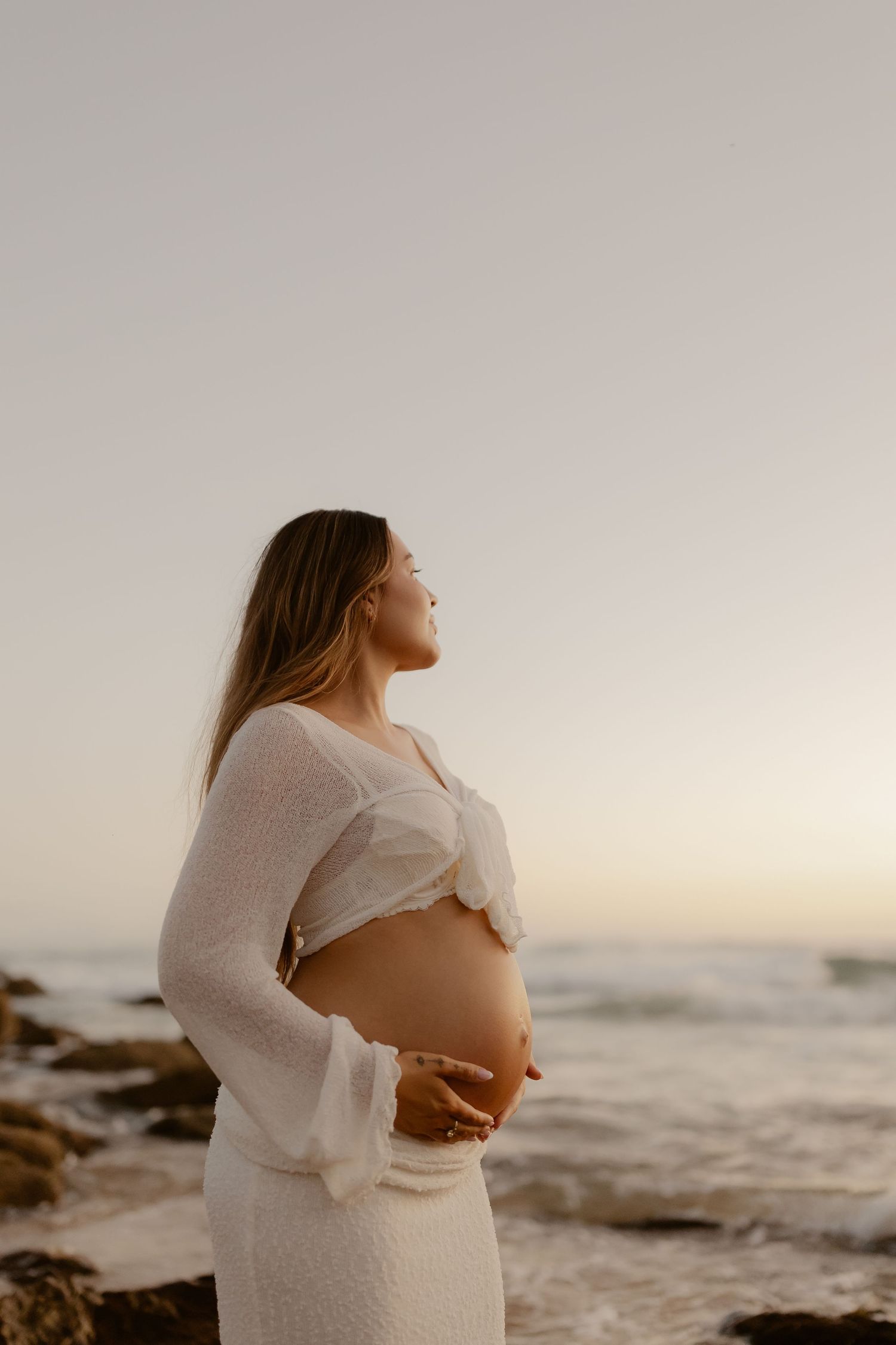 Maternity photos on beach at sunset showing woman in white dress cradling her belly.