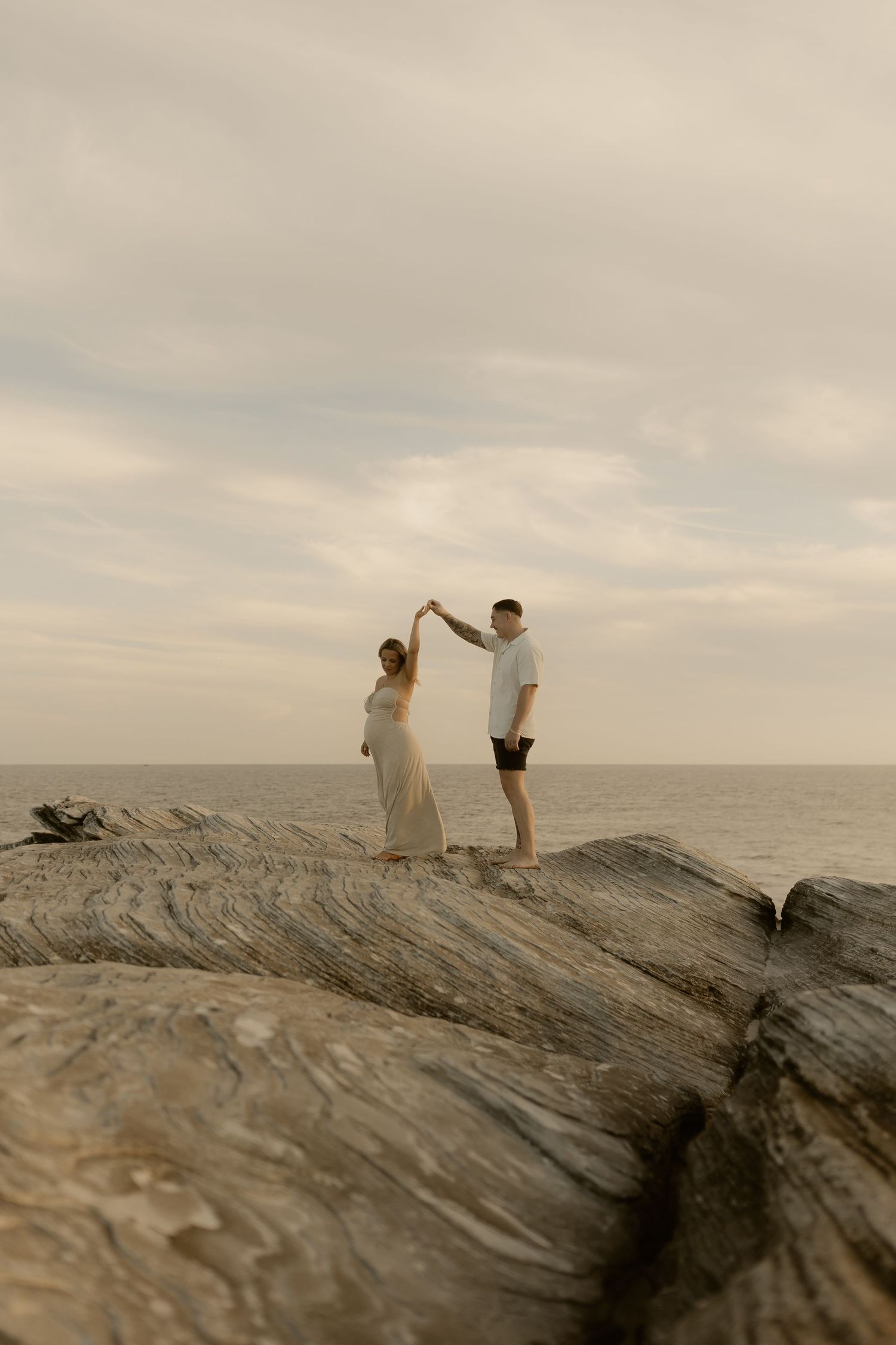 Two figures stand on a rocky coastal outcrop against a cloudy sky at sunset.