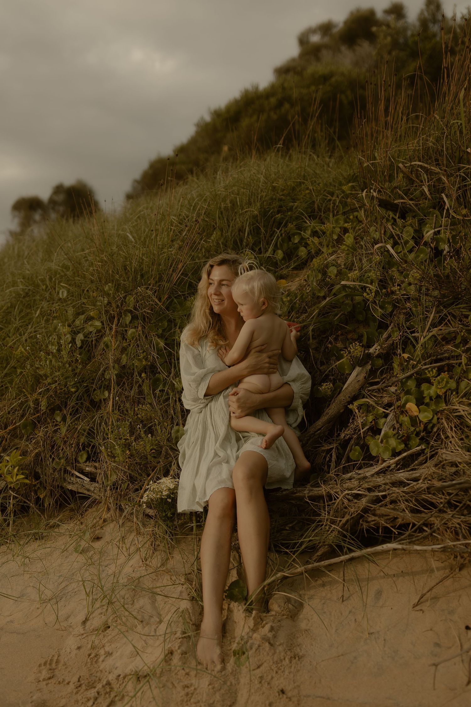 A tender moment captured on sandy dunes with green hillside vegetation in moody lighting.