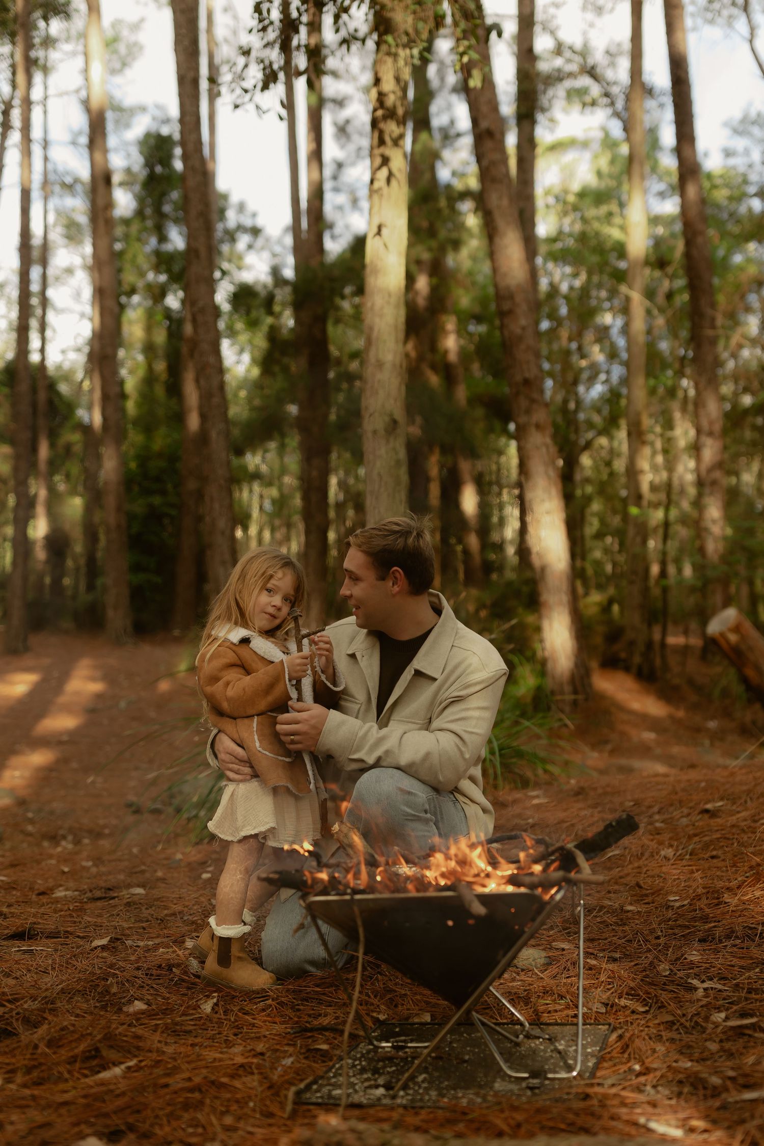 Father crouches with young daughter beside a small campfire in a pine forest.