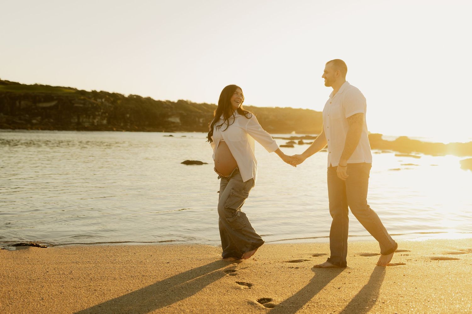 Pregnant couple holding hands walking on a sandy beach at golden sunset with footprints in the sand.