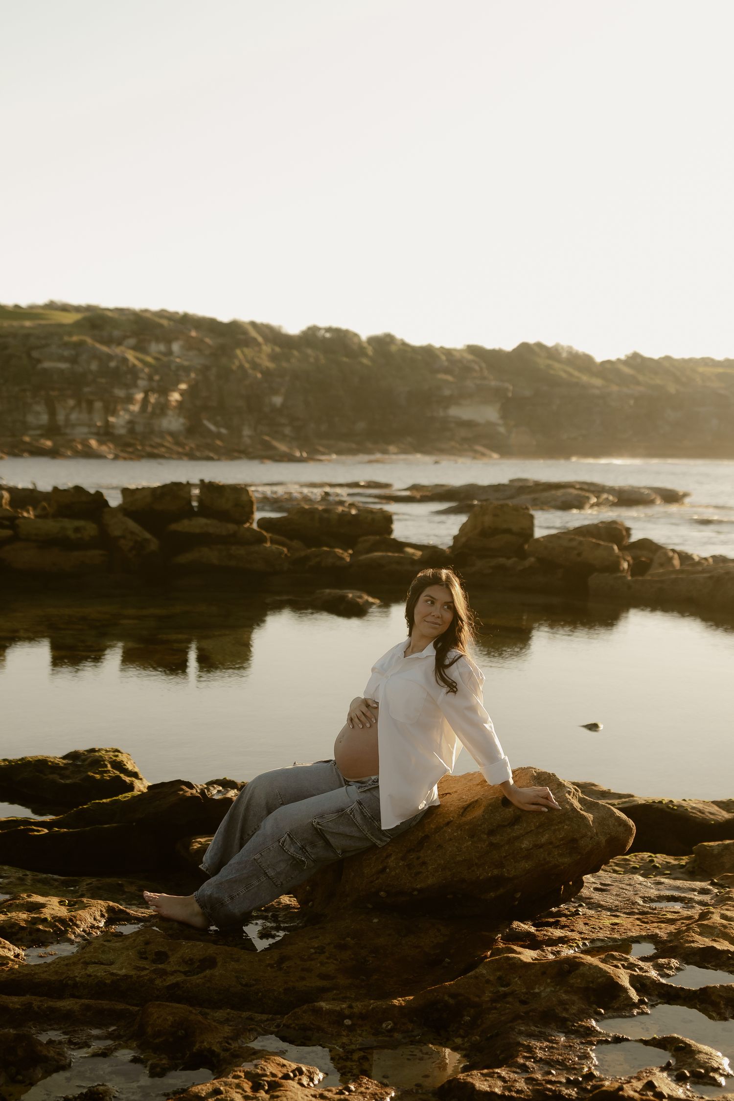 Pregnant woman in white shirt sits on coastal rocks at golden hour with calm ocean and cliffs behind her.