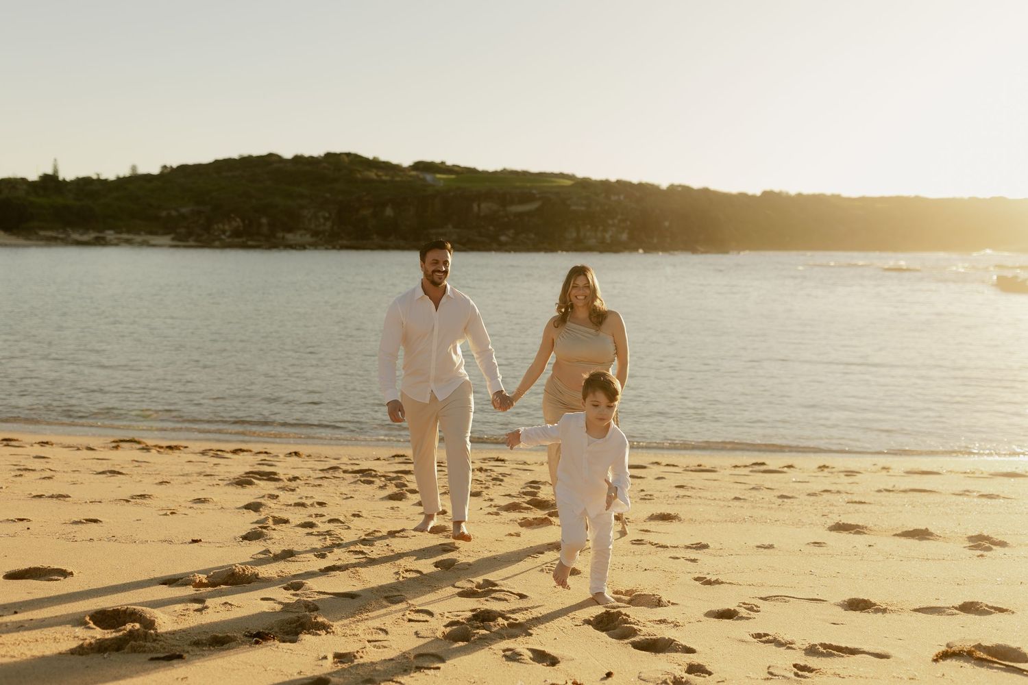 Family of three holding hands walking on a sunny beach at golden hour with calm water in the background.