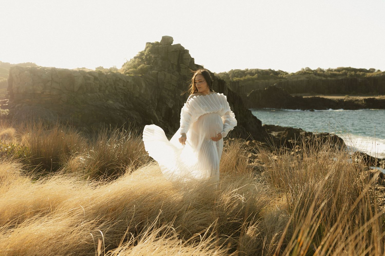 Woman in flowing white dress stands among tall grass near coastal cliffs at golden hour.
