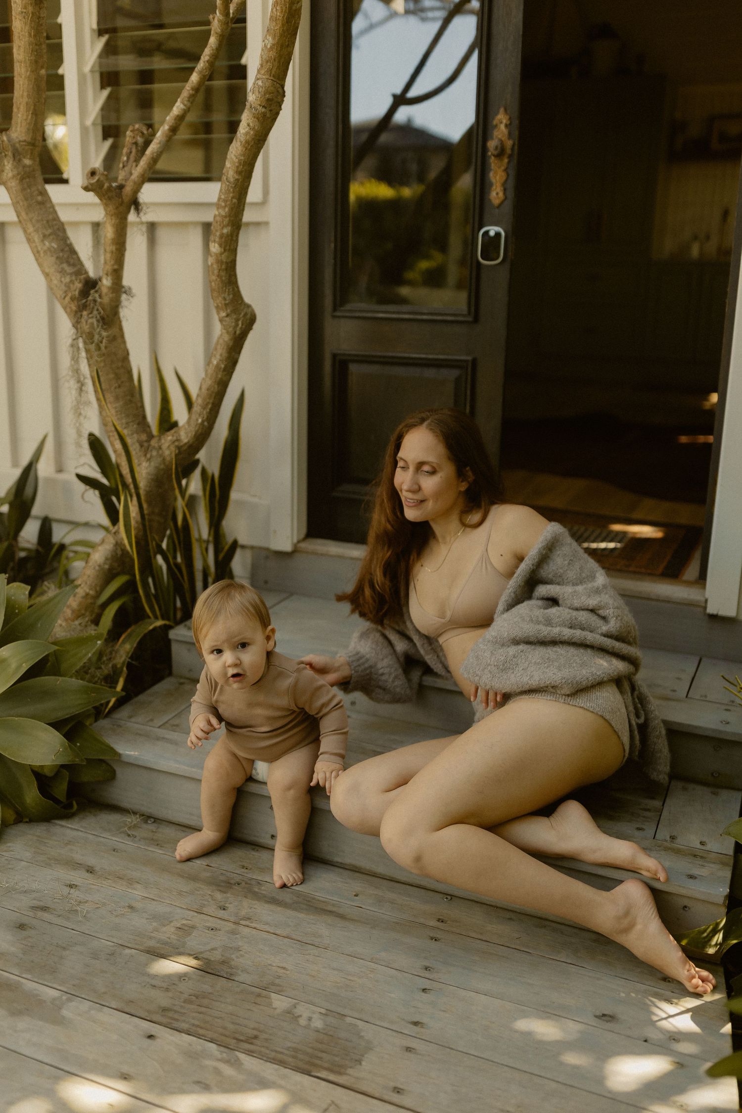 Adult and baby relaxing on sunny porch with tropical plants in background.