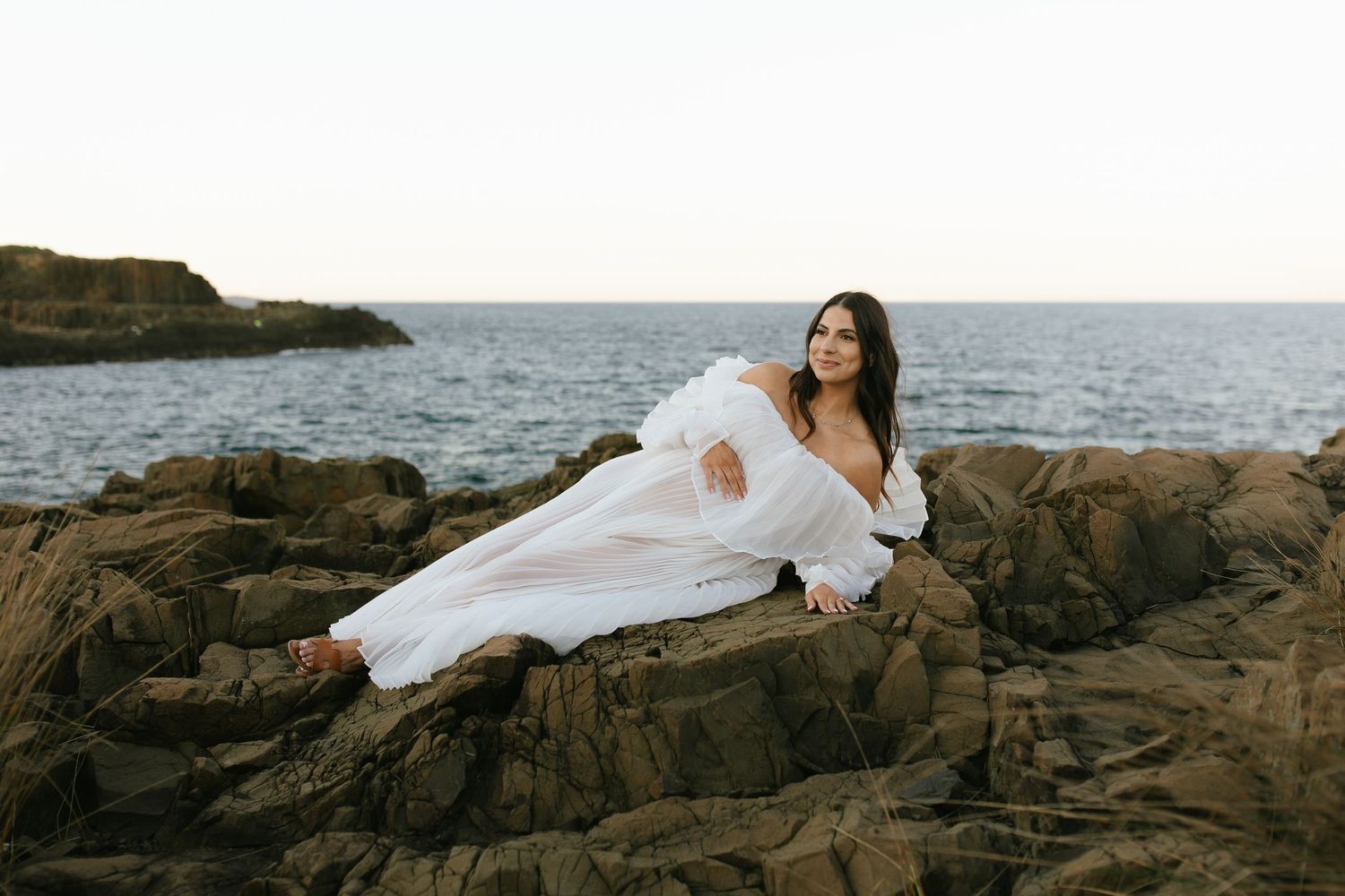 Person in white dress poses gracefully on rocky coastal shoreline with ocean waves in background.