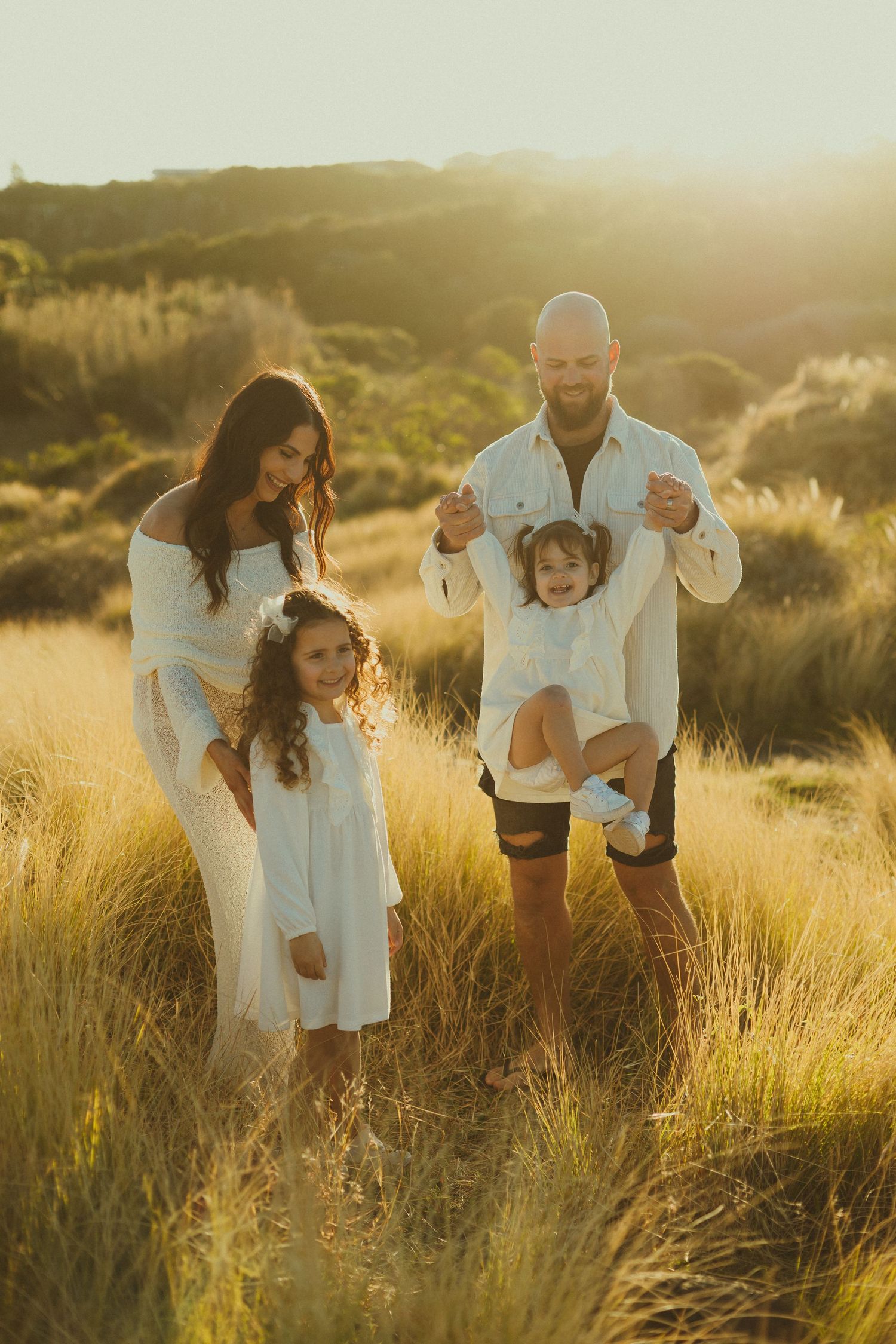 Family in coordinated white outfits share joyful moment in sun-drenched field at golden hour.