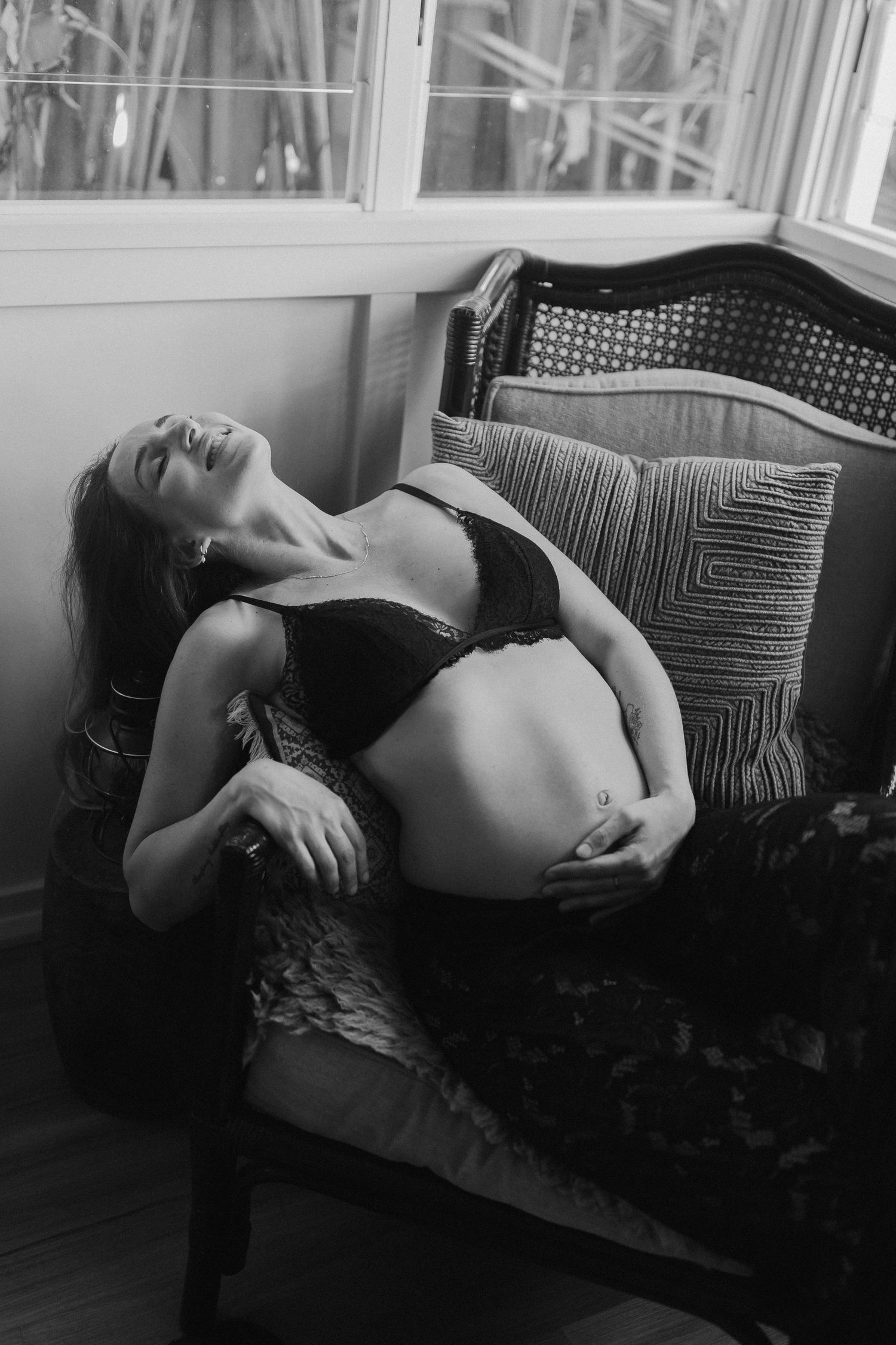 Artistic black and white maternity photo of an expectant mother reclining on a vintage couch by a window.