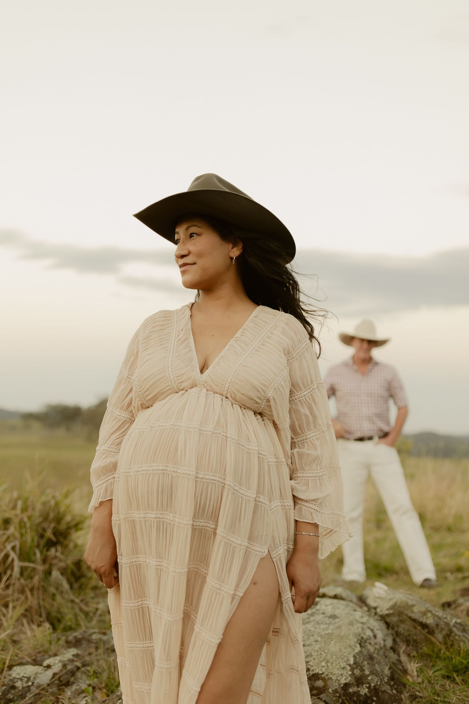 Pregnant woman in beige lace dress and cowboy hat stands in field with man in background at dusk.