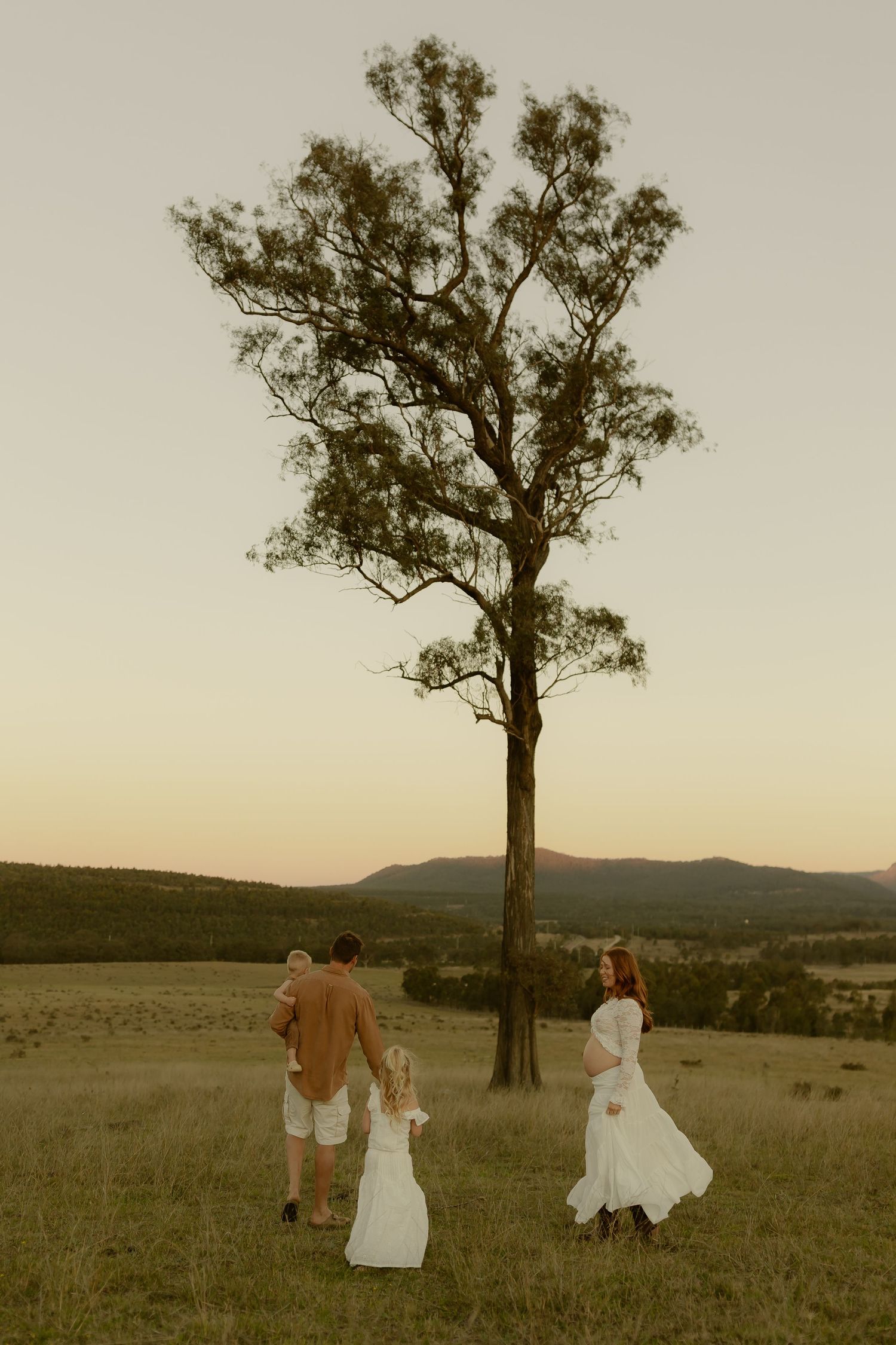 A family stands near a tall tree in an open field at dusk, with mountains in the background.