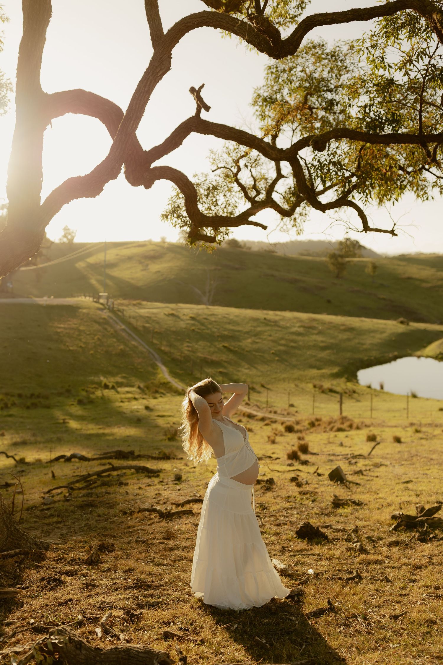 Pregnant woman in white dress standing under sprawling oak tree at golden sunset on hillside.