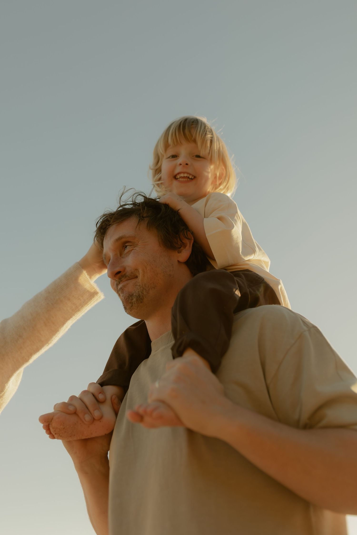 A father carries his laughing blonde toddler on his shoulders against a clear sky in warm golden sunlight.