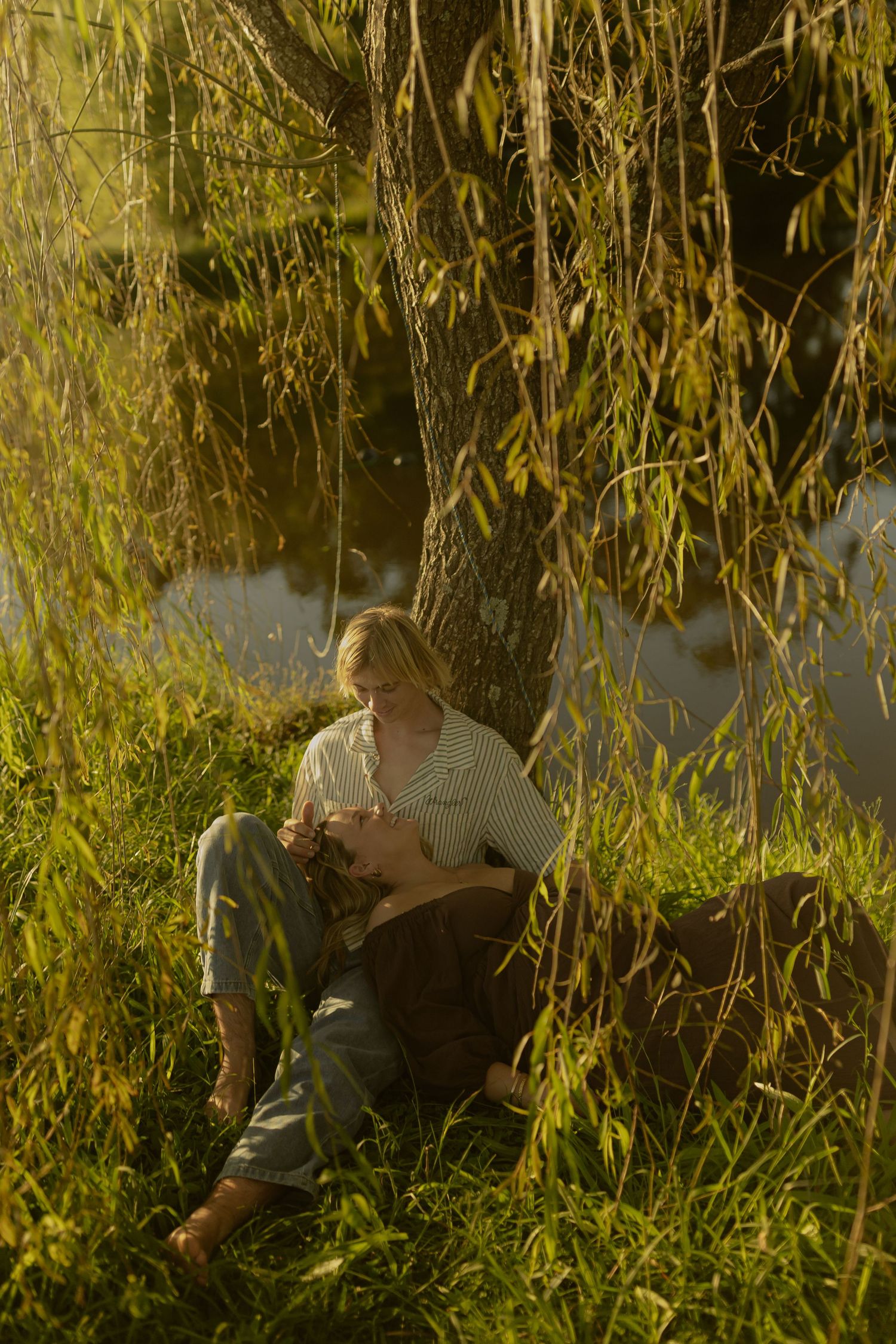 Person sitting under a weeping willow tree by a pond in golden sunlight, surrounded by lush green grass.