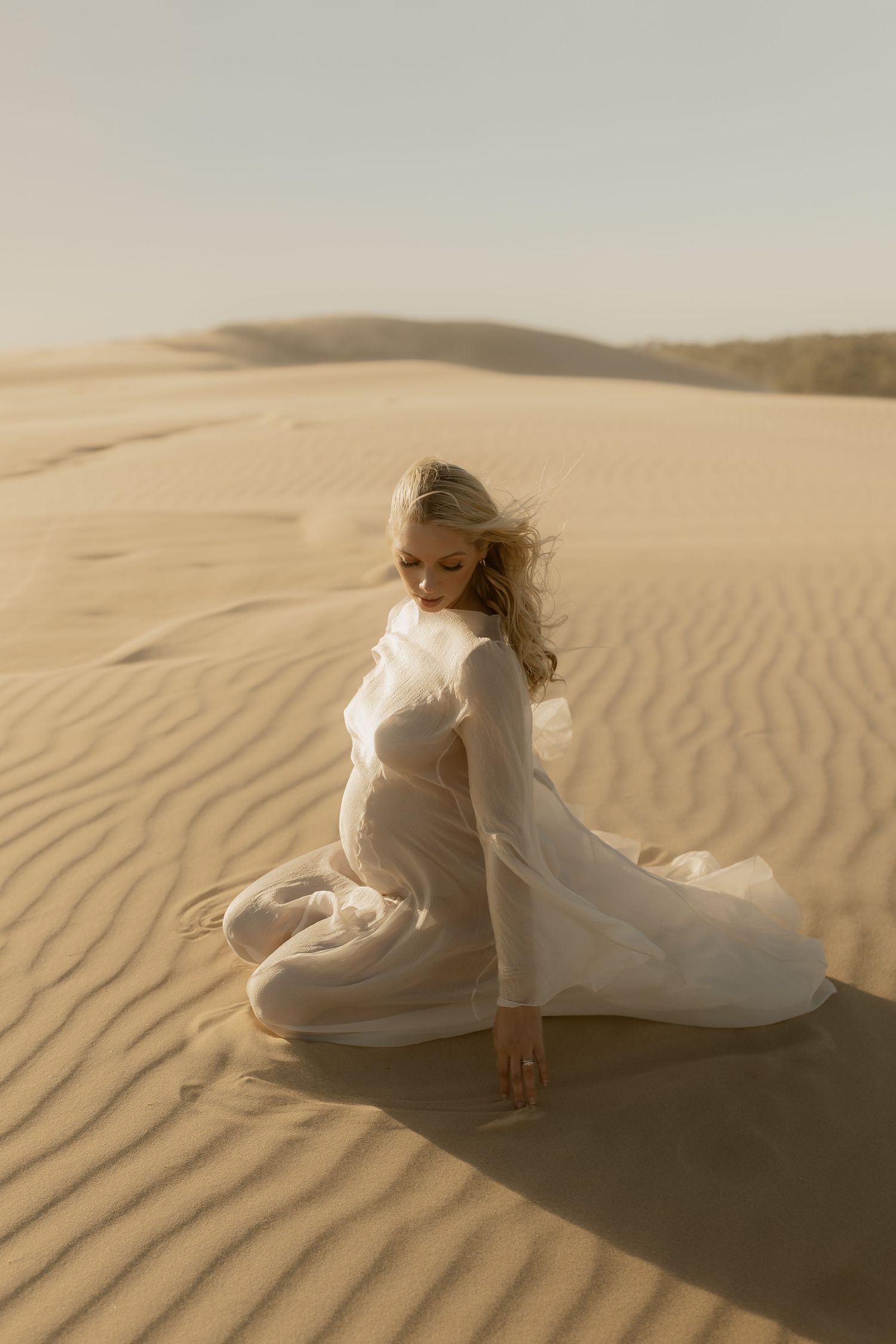 Pregnant woman in white sheer gown kneeling gracefully on rippled desert sand dunes.