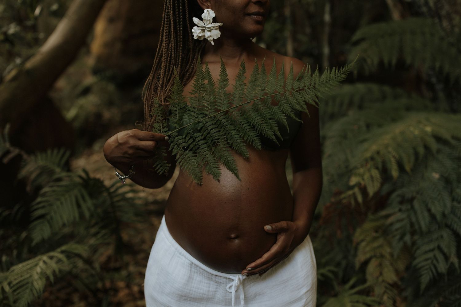 Pregnant Black woman holding fern leaf in tropical forest, wearing white skirt with flower hair accessory.