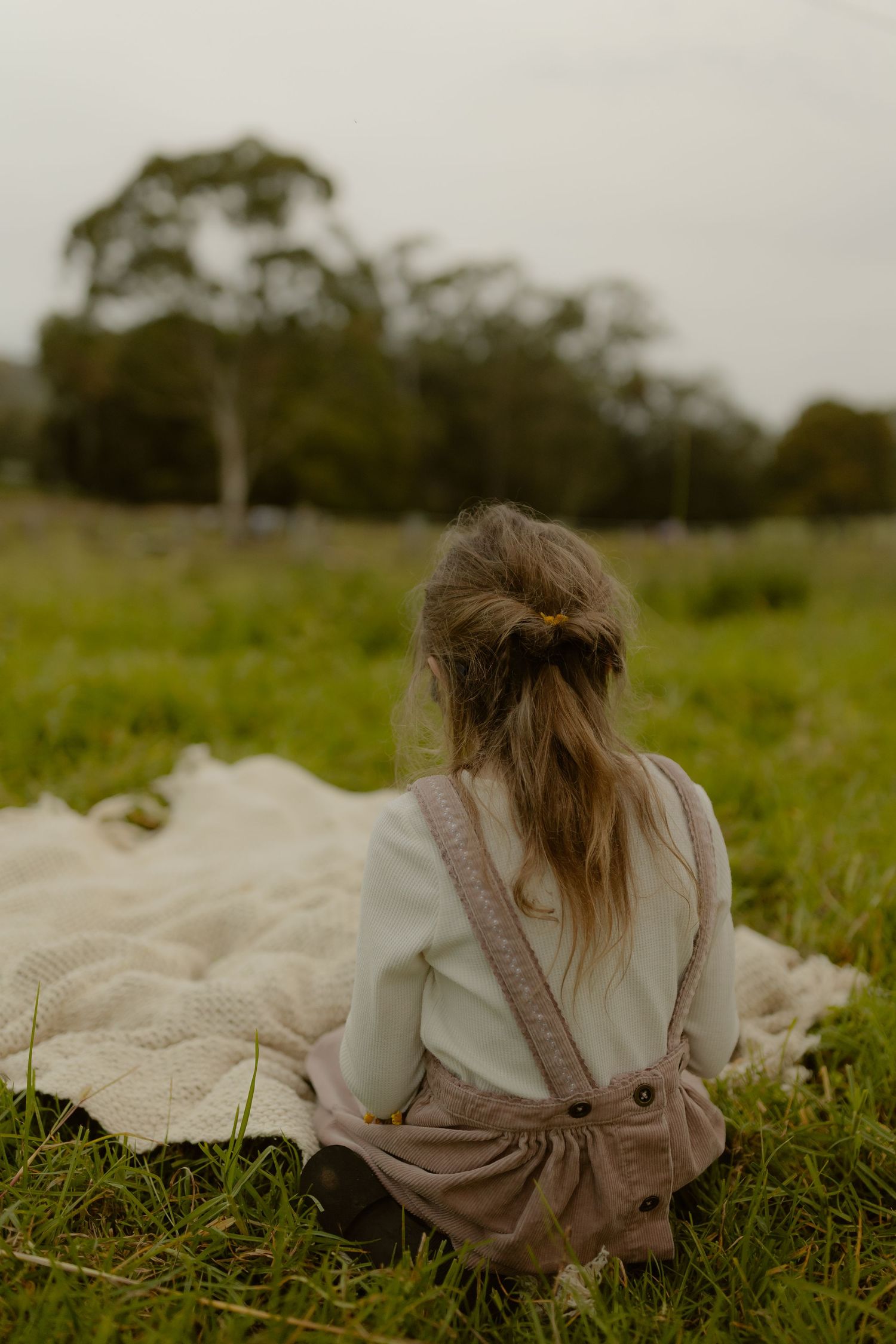 Young girl in pink overalls sits on a white blanket in a lush green field, viewed from behind.