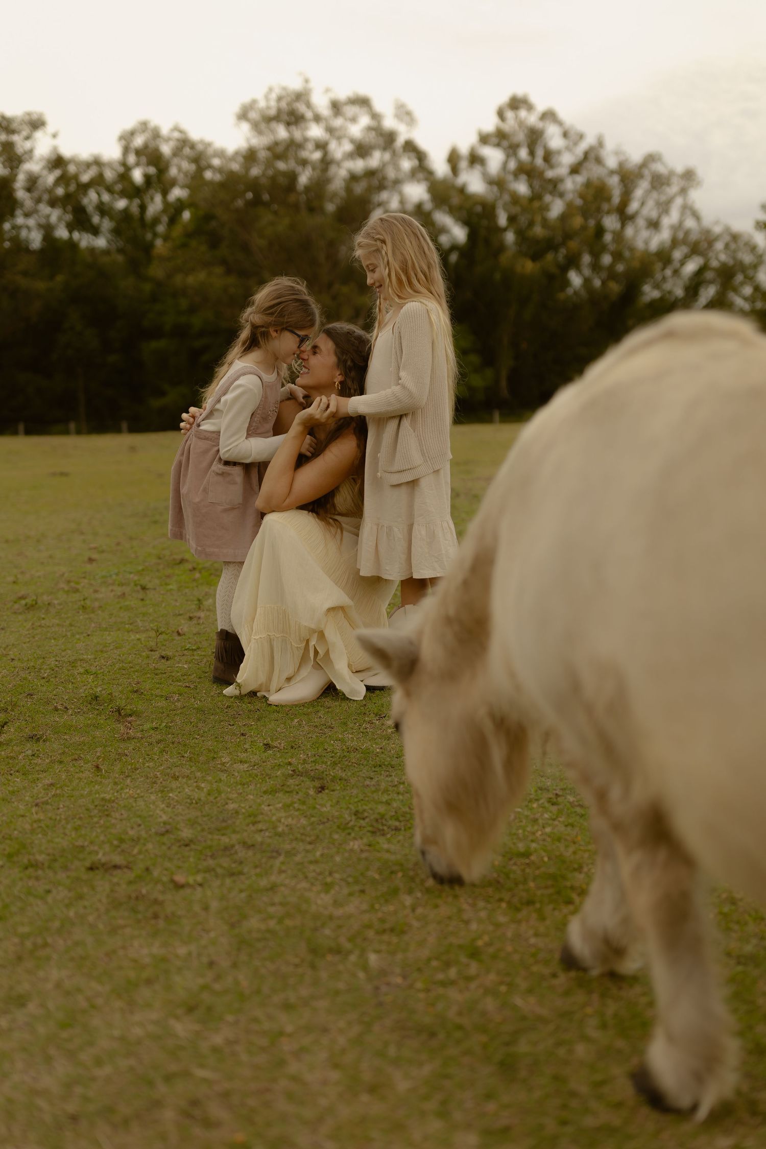 Mother kneeling in cream dress embracing two young girls on a green farm with a white cow nearby.