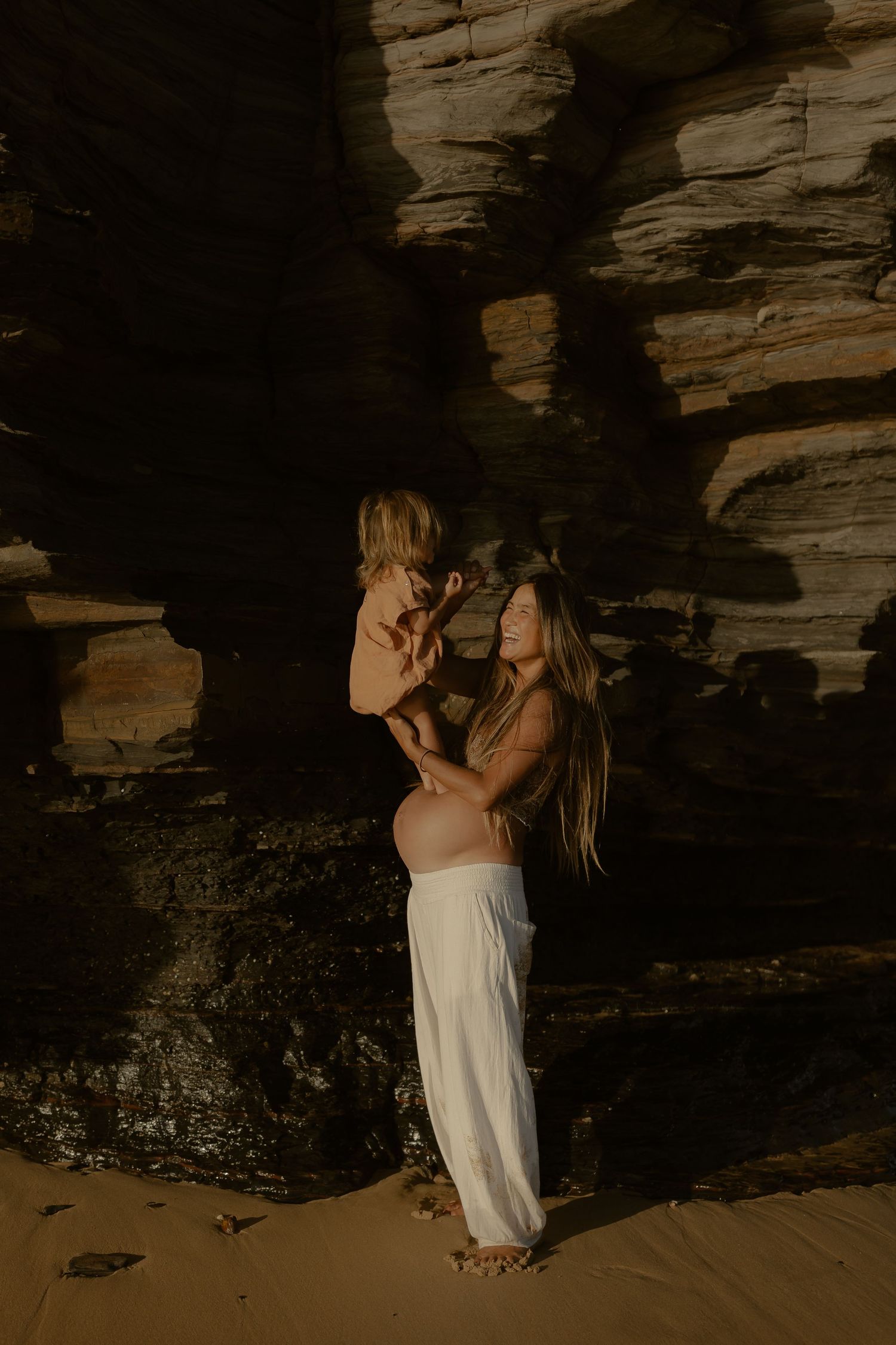 Pregnant woman in white skirt lifts toddler near rocky cliffs on sandy beach in warm golden light.