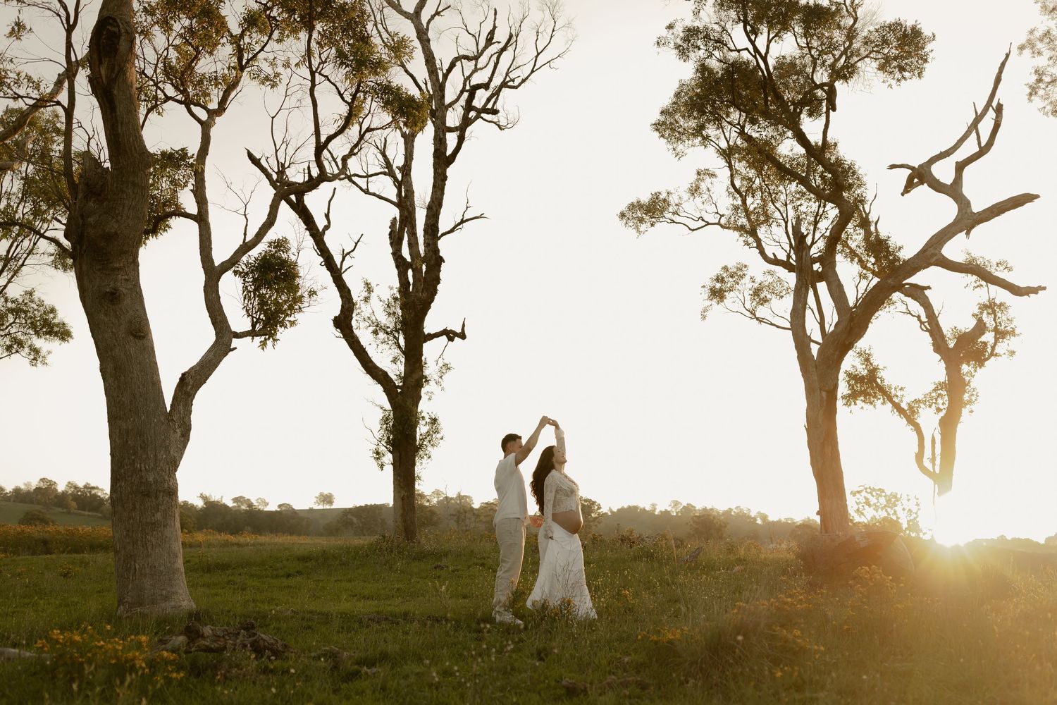 Couple dancing at sunset among gum trees in a lush green field, golden light glowing behind them.