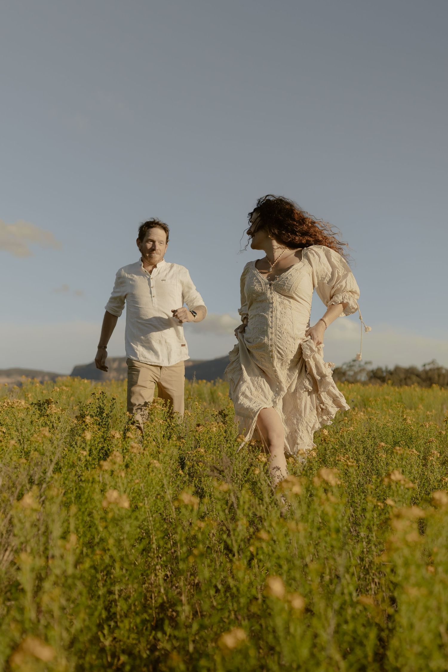 A couple in light colored outfits runs playfully through a wildflower meadow at sunset.