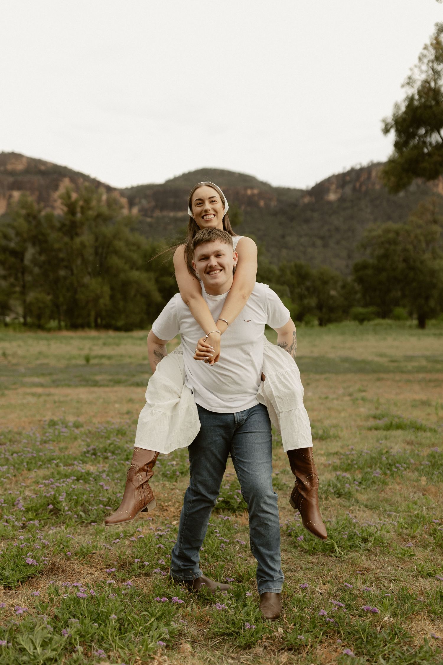 Playful couple shares joyful moments in a grassy field with mountains behind them.