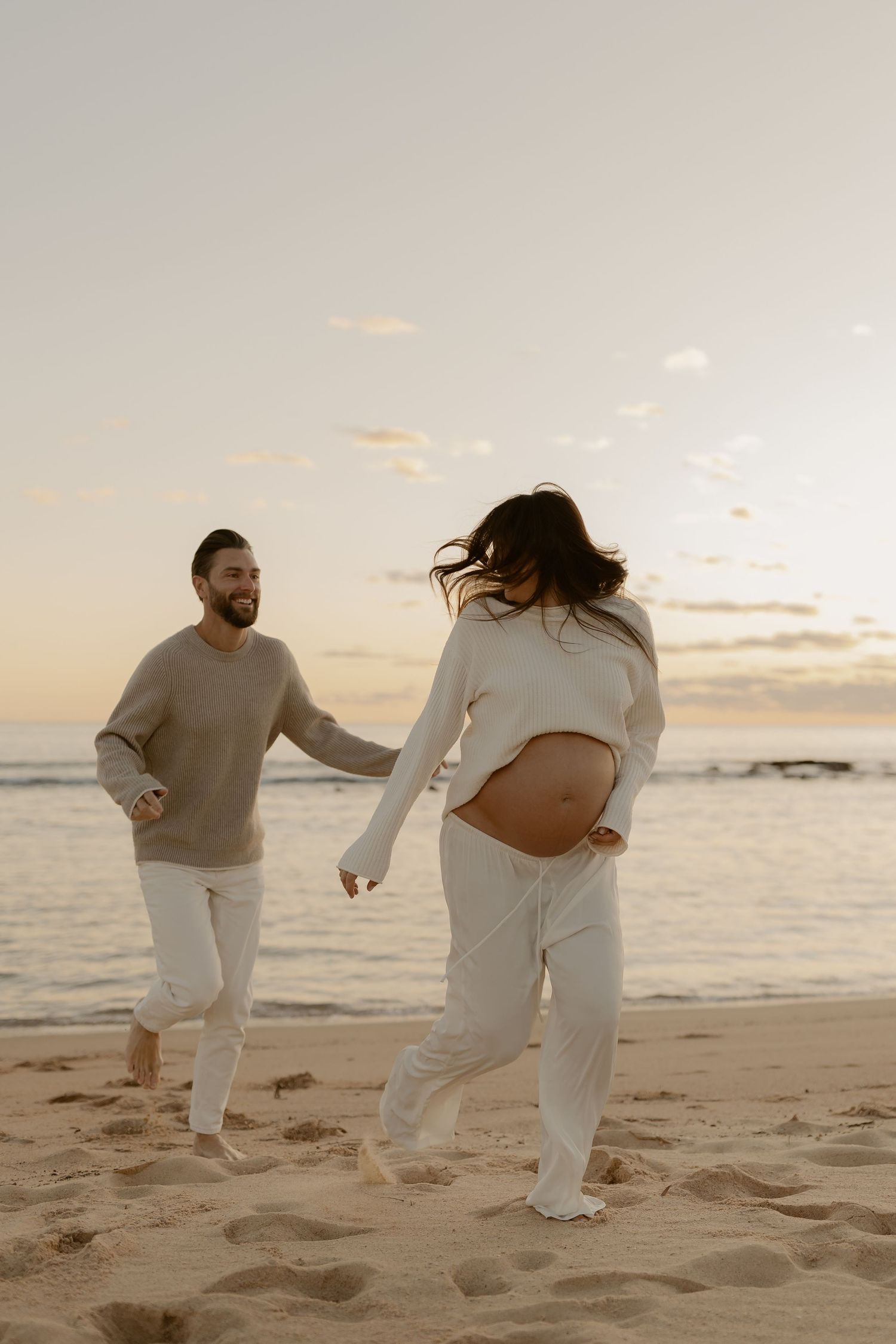 Couple in matching white outfits playfully running and dancing together on a beach at sunset.