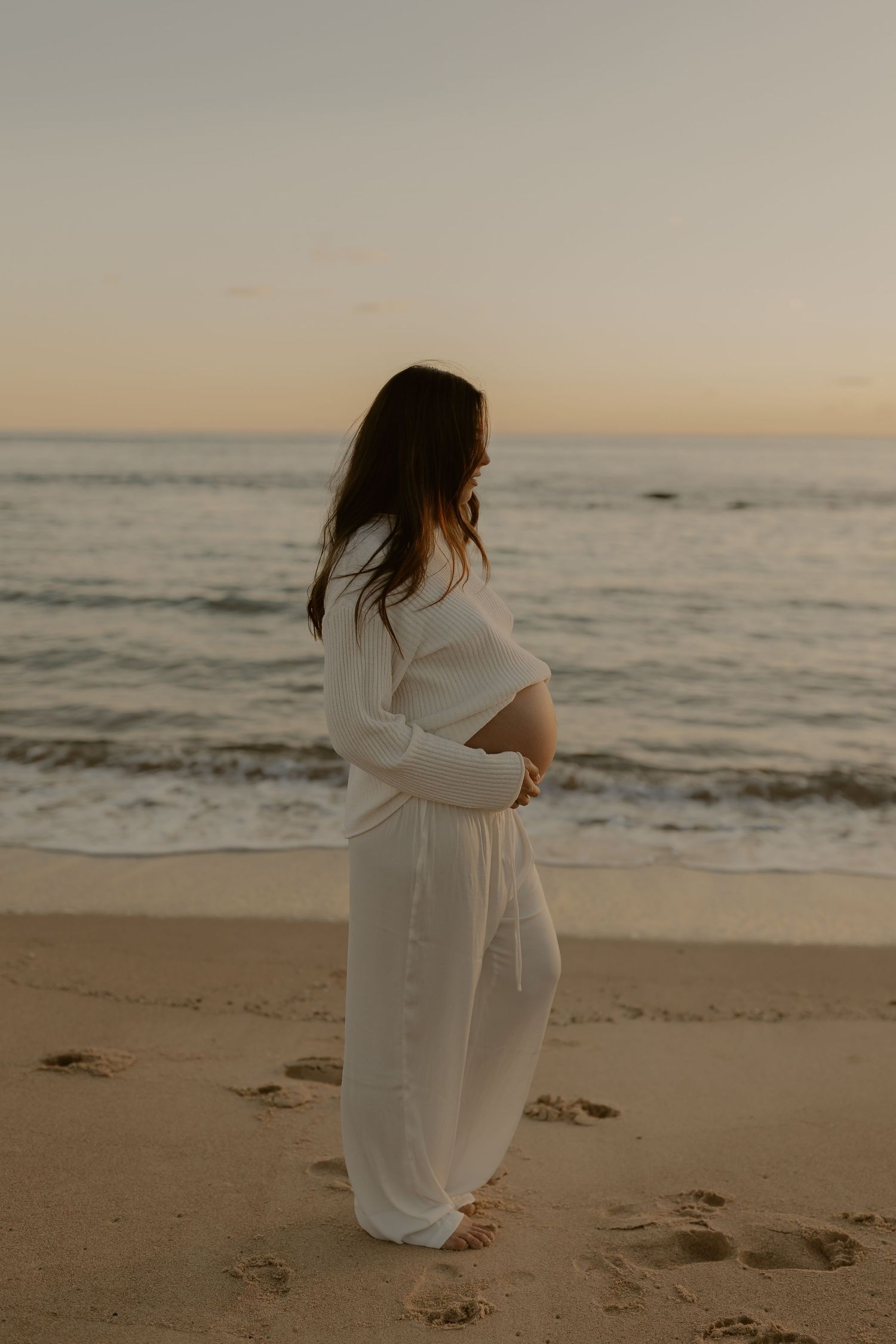Silhouette of a pregnant woman in white dress walking along a beach at sunset.