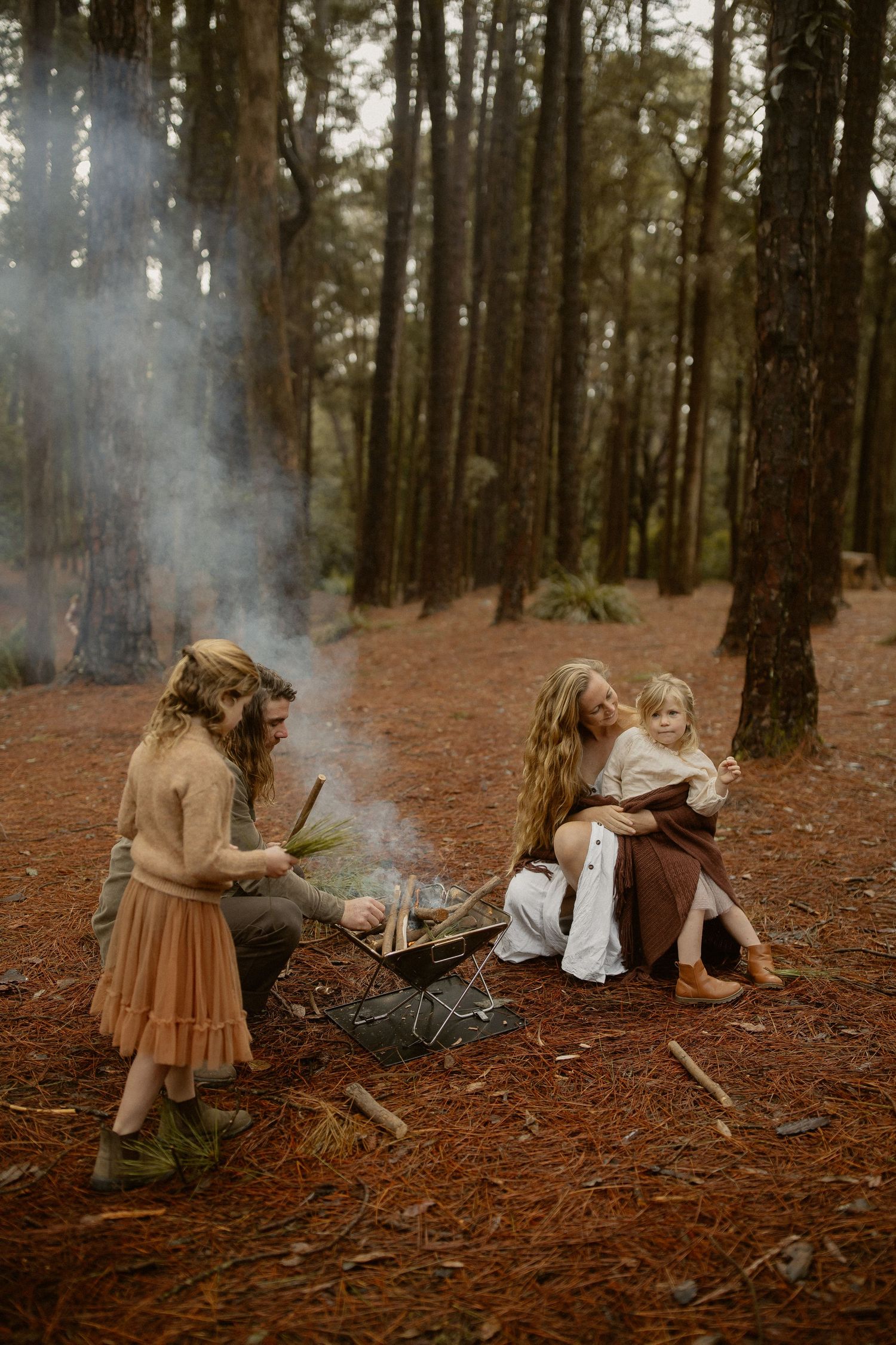 A moody forest scene with smoke and vintage-styled figures sitting near a campfire on pine needle covered ground.