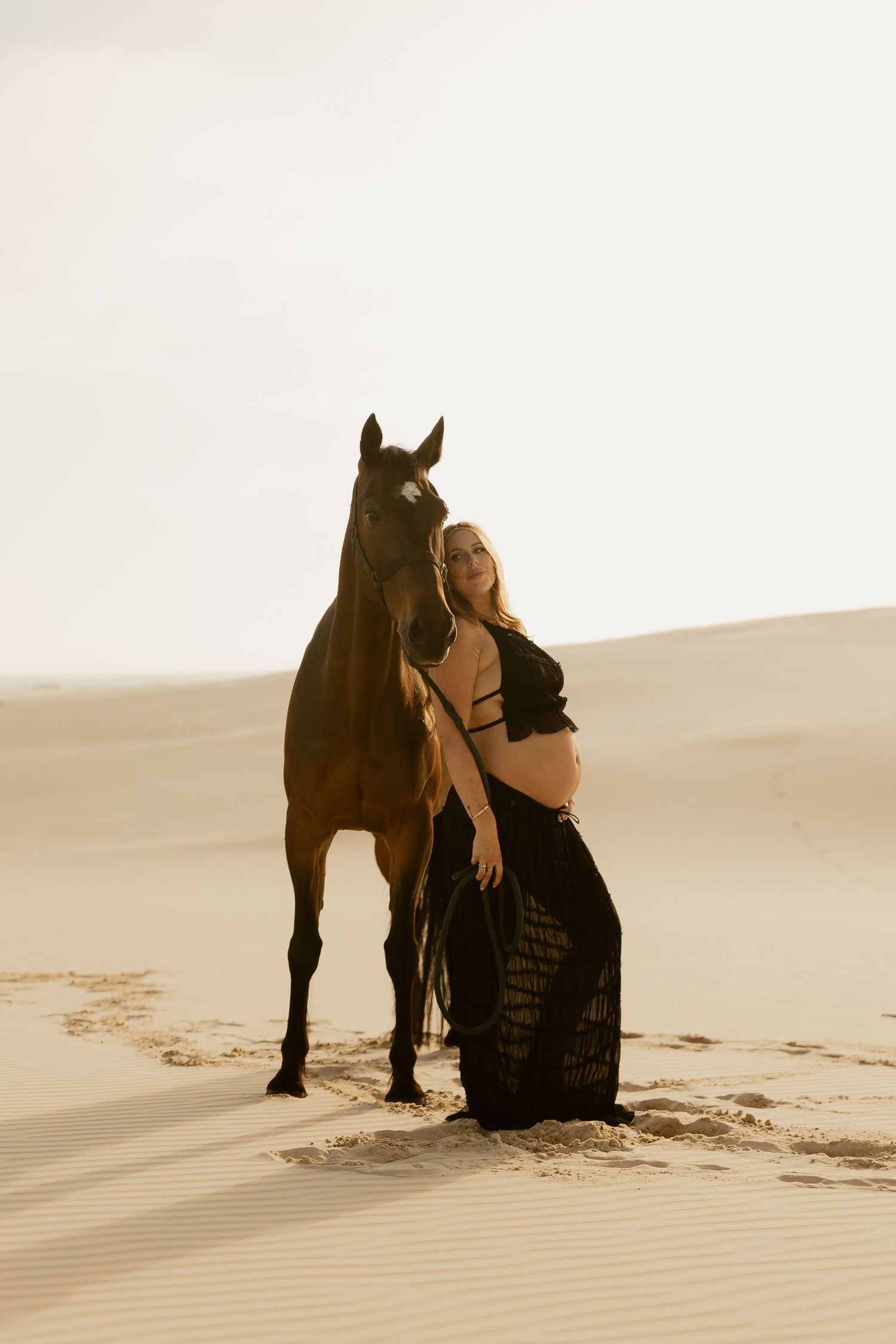 A figure in a black dress poses with a horse against sand dunes in a sepia-toned artistic photoshoot.