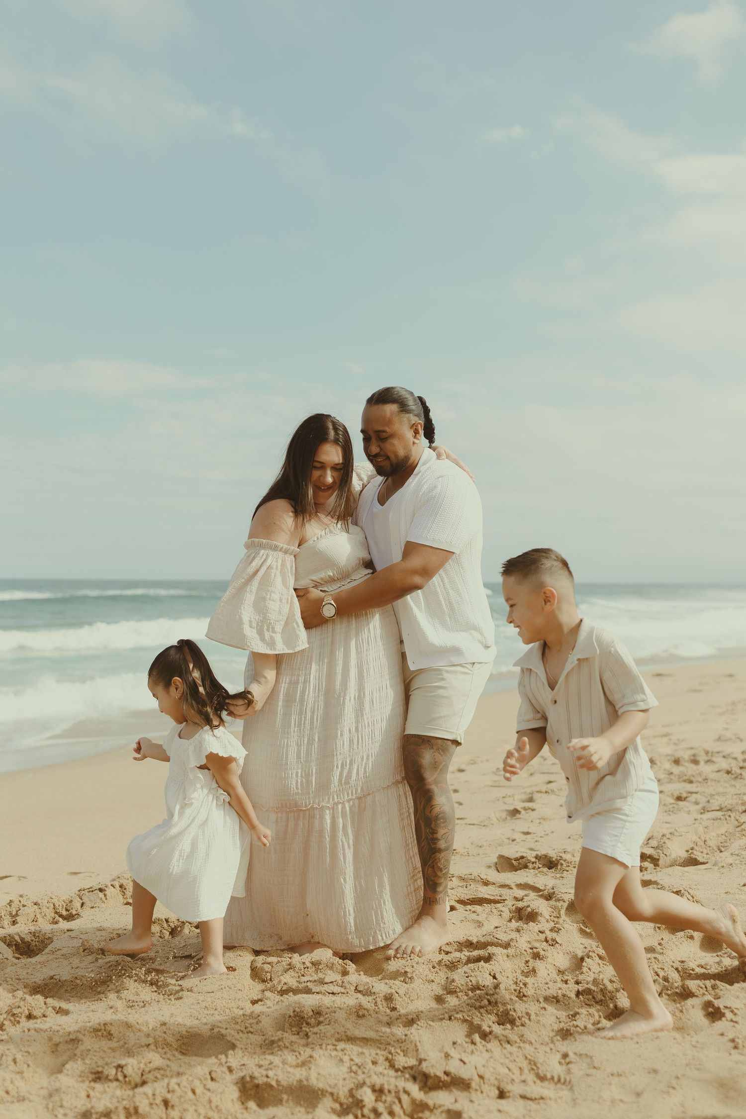 A family wearing white outfits plays together on a sandy beach with ocean waves in the background.
