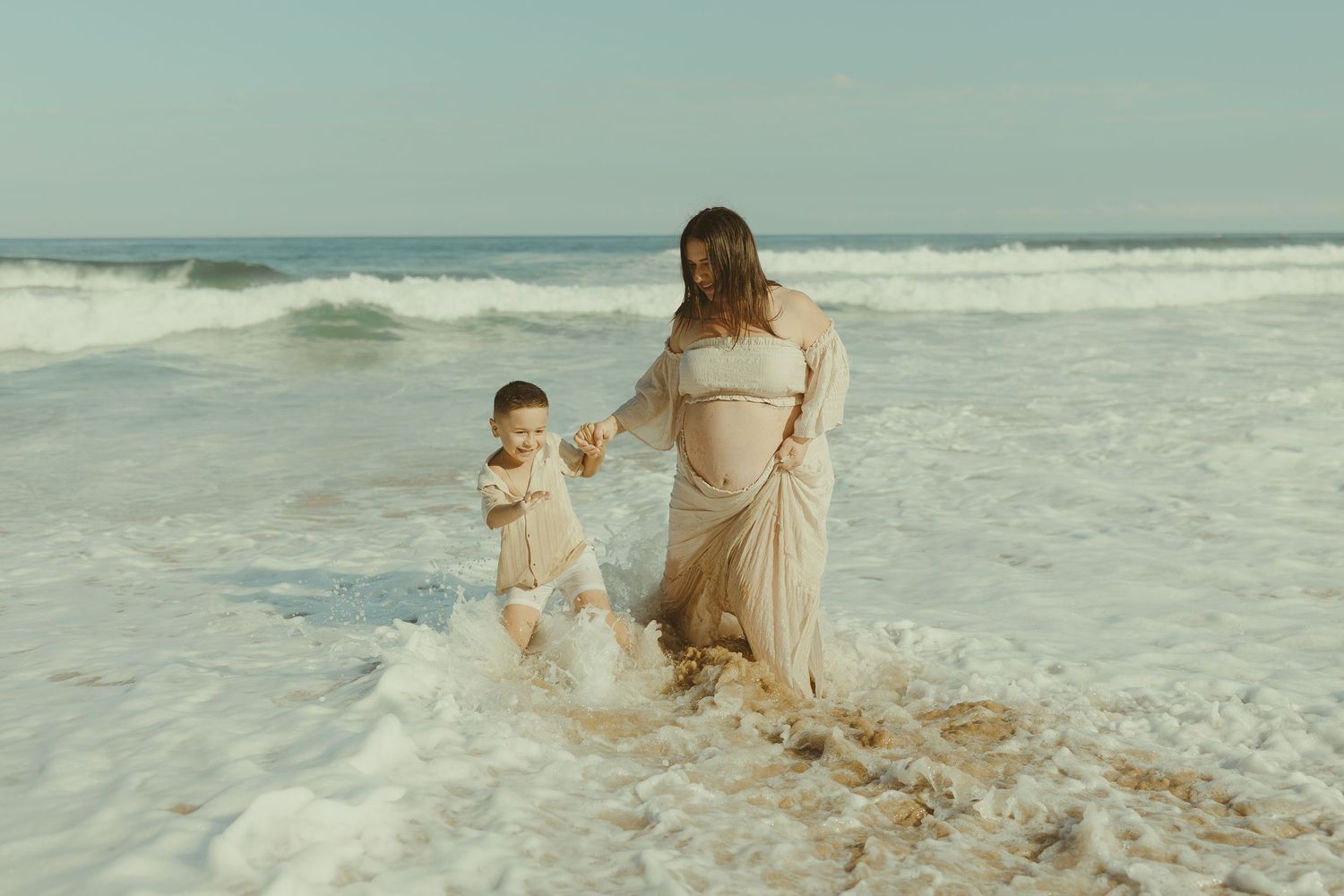 People in flowy outfits walk along a scenic beach with crashing waves and soft sand during golden hour.